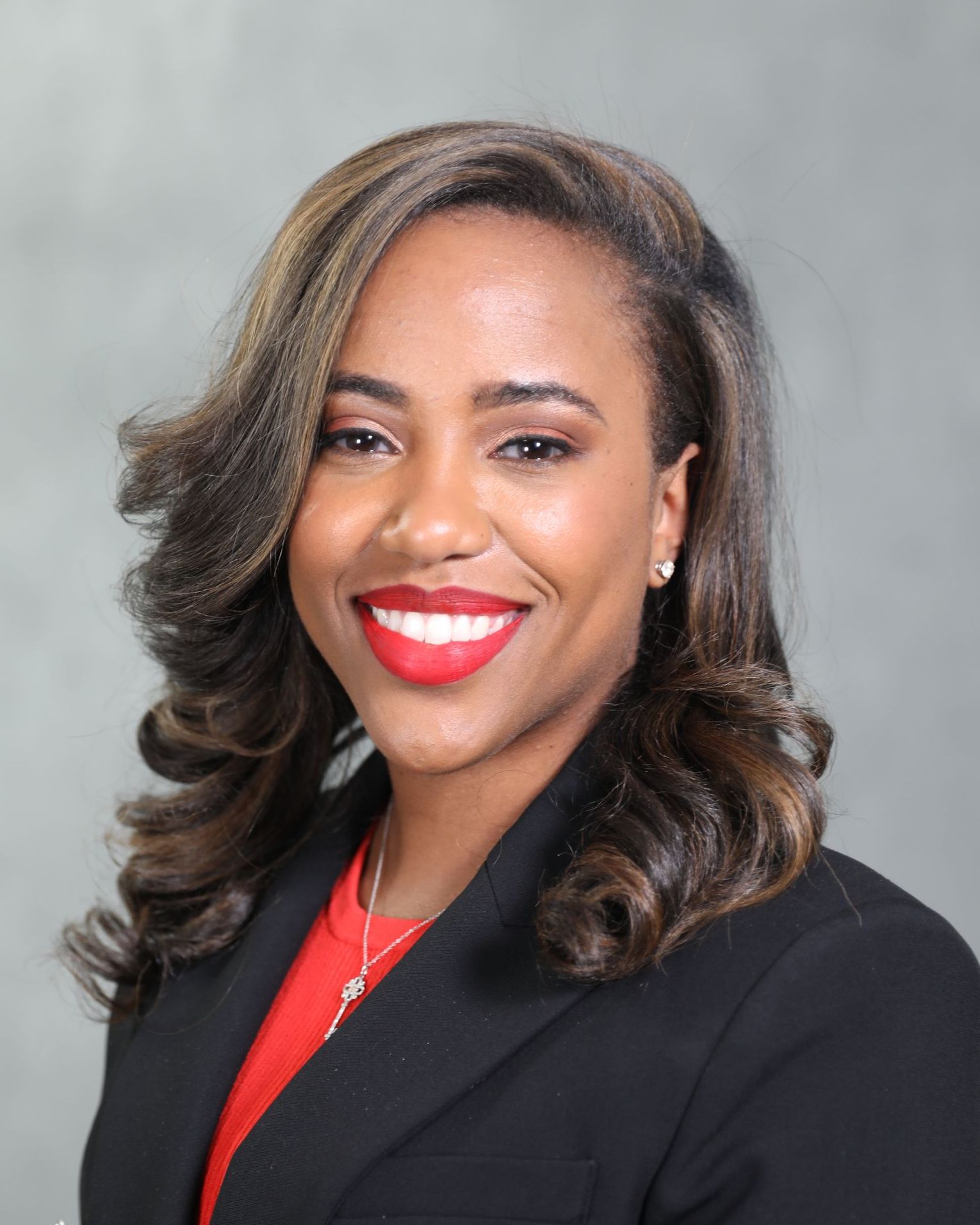 A professional headshot of a smiling woman wearing a red top and a black blazer against a grey background.