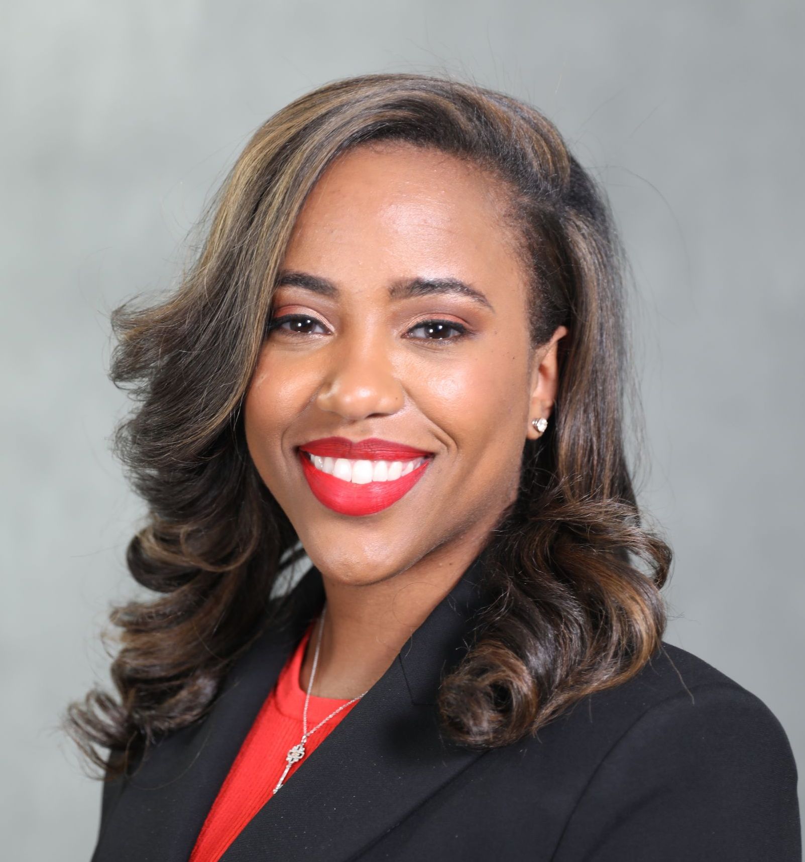 A smiling person wearing a black blazer over a red top, set against a plain gray background.