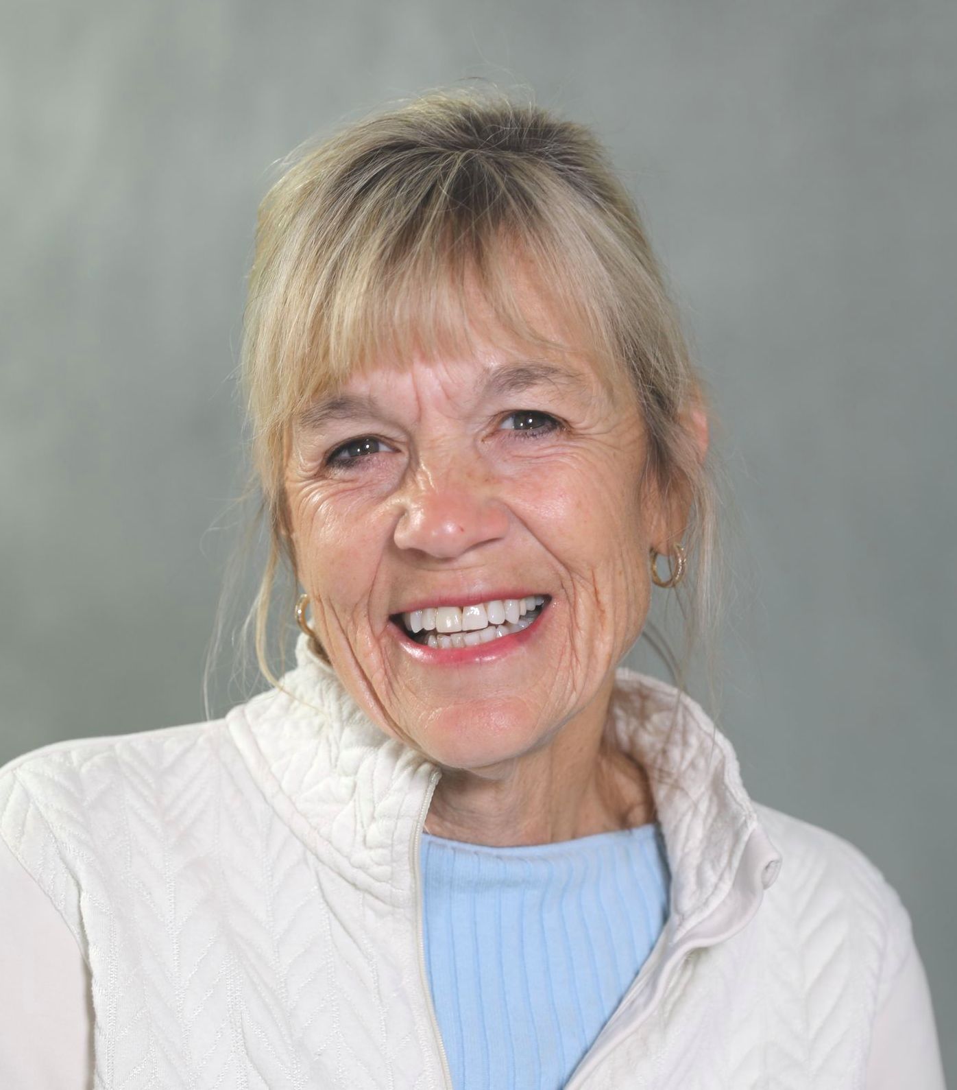 A smiling person with blonde hair wearing a white jacket over a light blue top against a gray background.