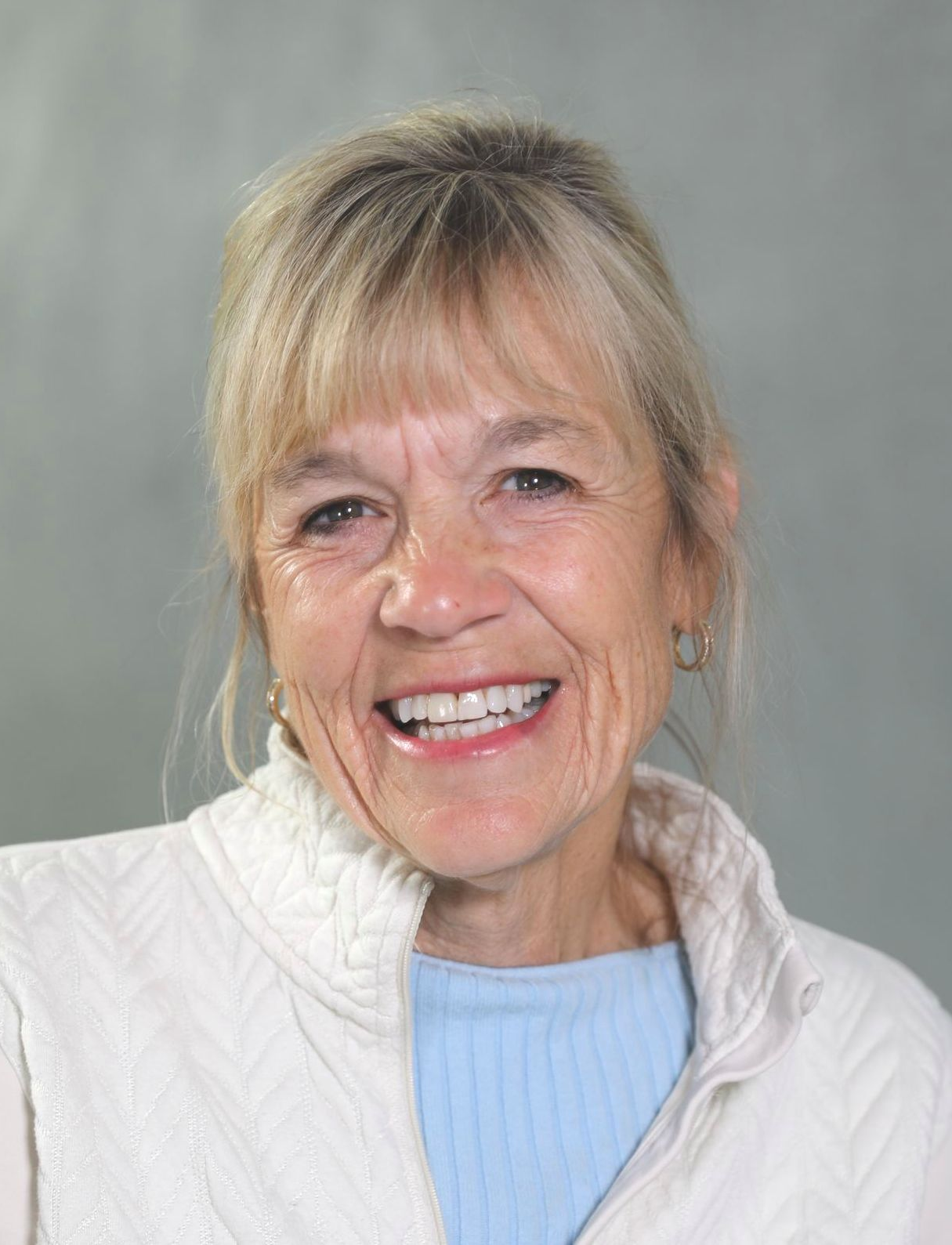 Smiling person with blonde hair wearing a light blue shirt under a white zip-up jacket against a grey background.