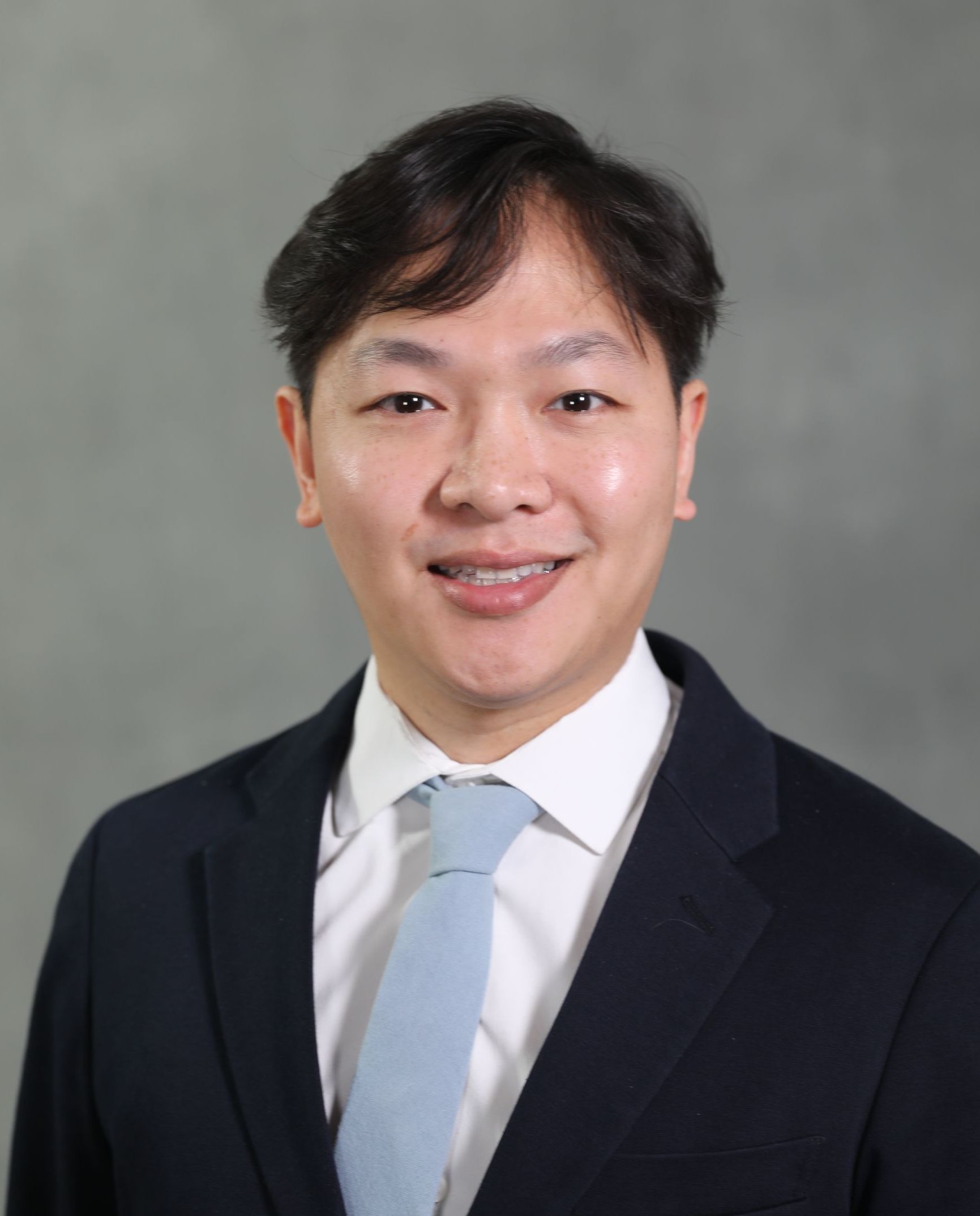 A headshot of a person wearing a dark blue suit, white shirt, and light blue tie against a gray background.