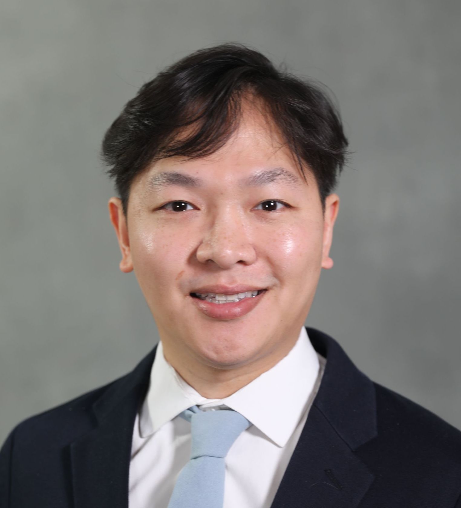 Headshot of a person wearing a dark blue suit, white shirt, and light blue tie against a neutral grey background.