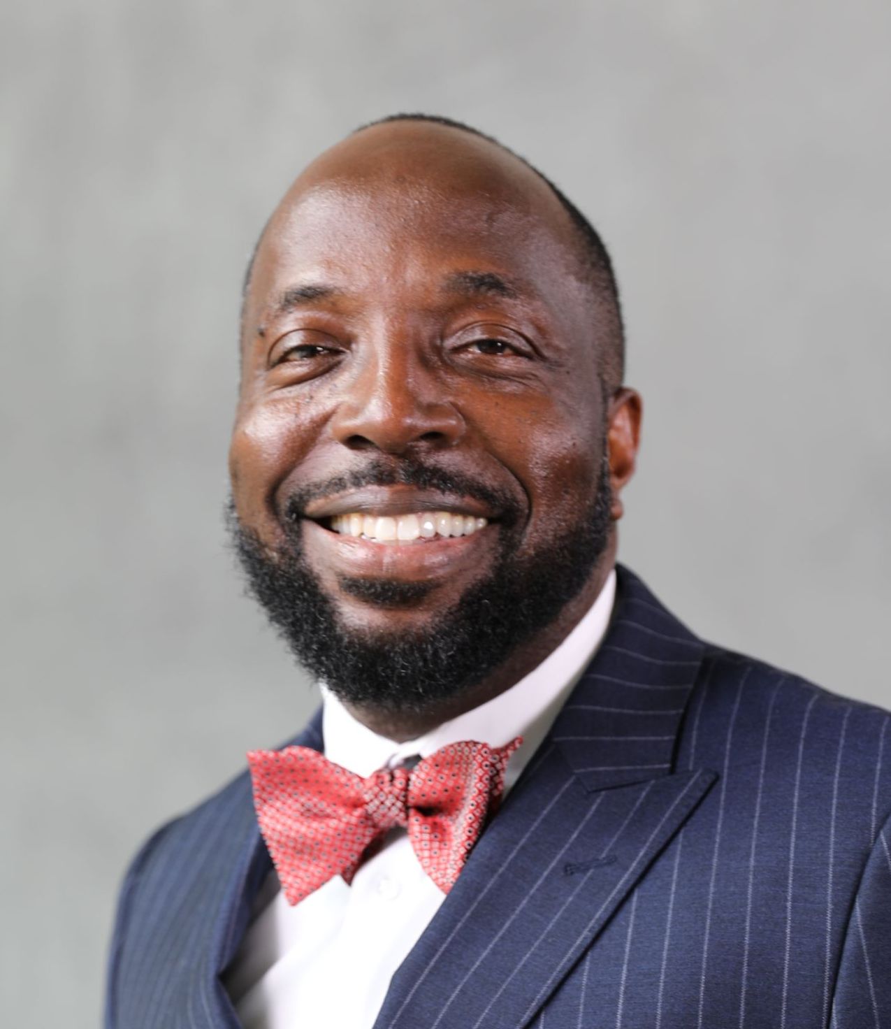 A person wearing a pinstripe suit and coral bowtie smiles warmly against a plain gray background.