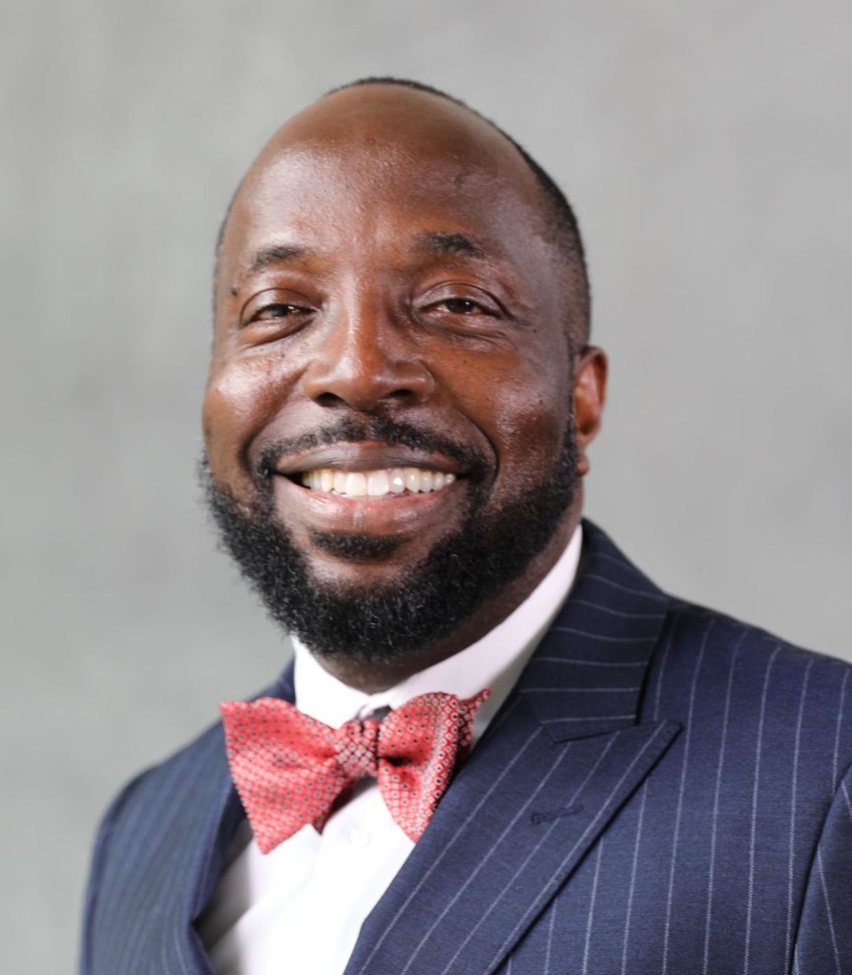 A person smiling, wearing a pinstripe navy suit, white collared shirt, and a coral bow tie against a gray background.