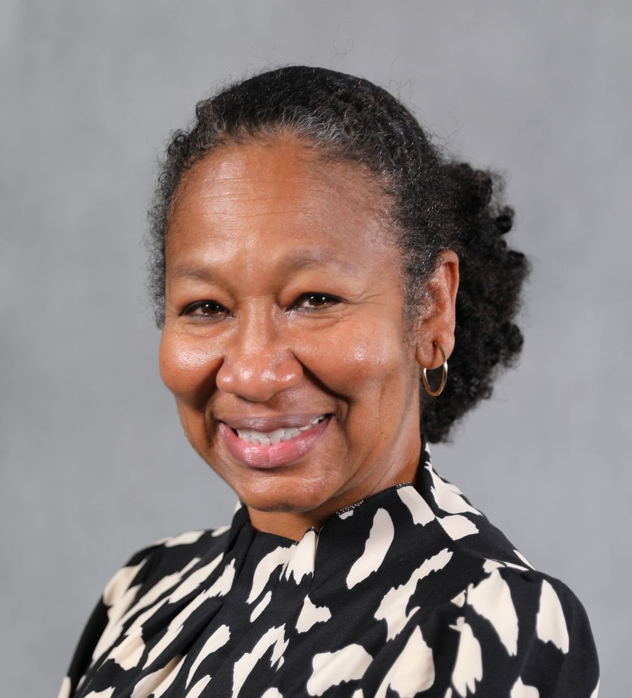 A smiling professional wearing a black and white patterned top against a plain gray background.