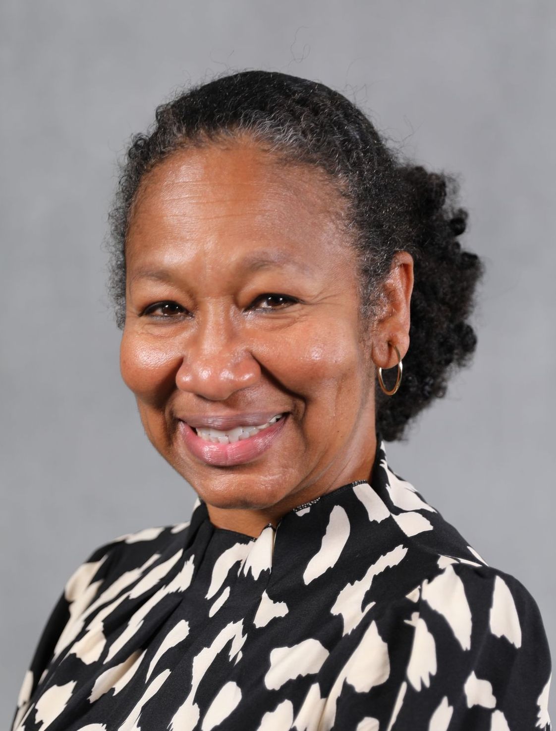 A smiling person with dark hair tied back, wearing a black and cream patterned blouse against a grey backdrop.