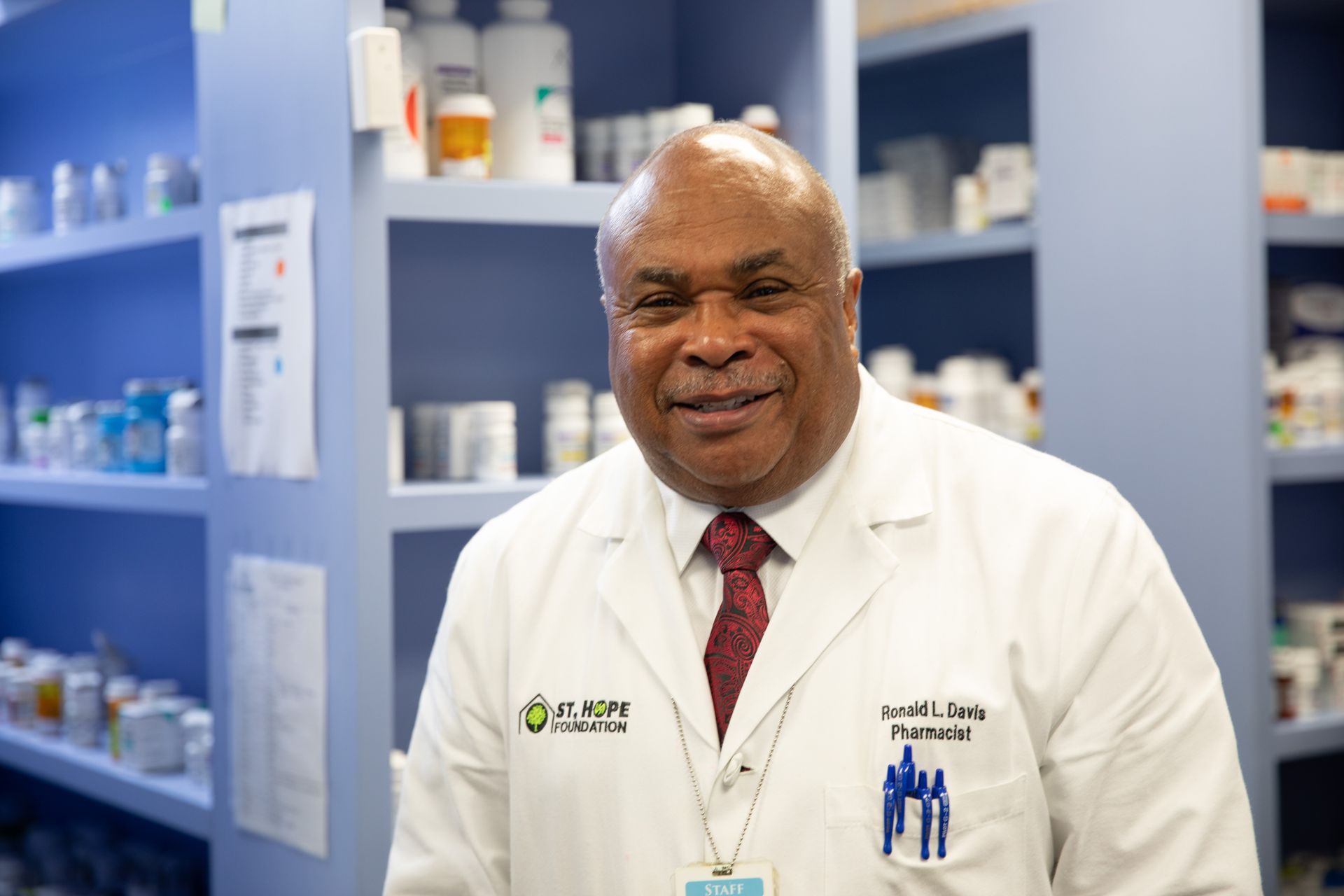 A smiling pharmacist in a white coat stands in front of blue pharmacy shelves filled with medication bottles.