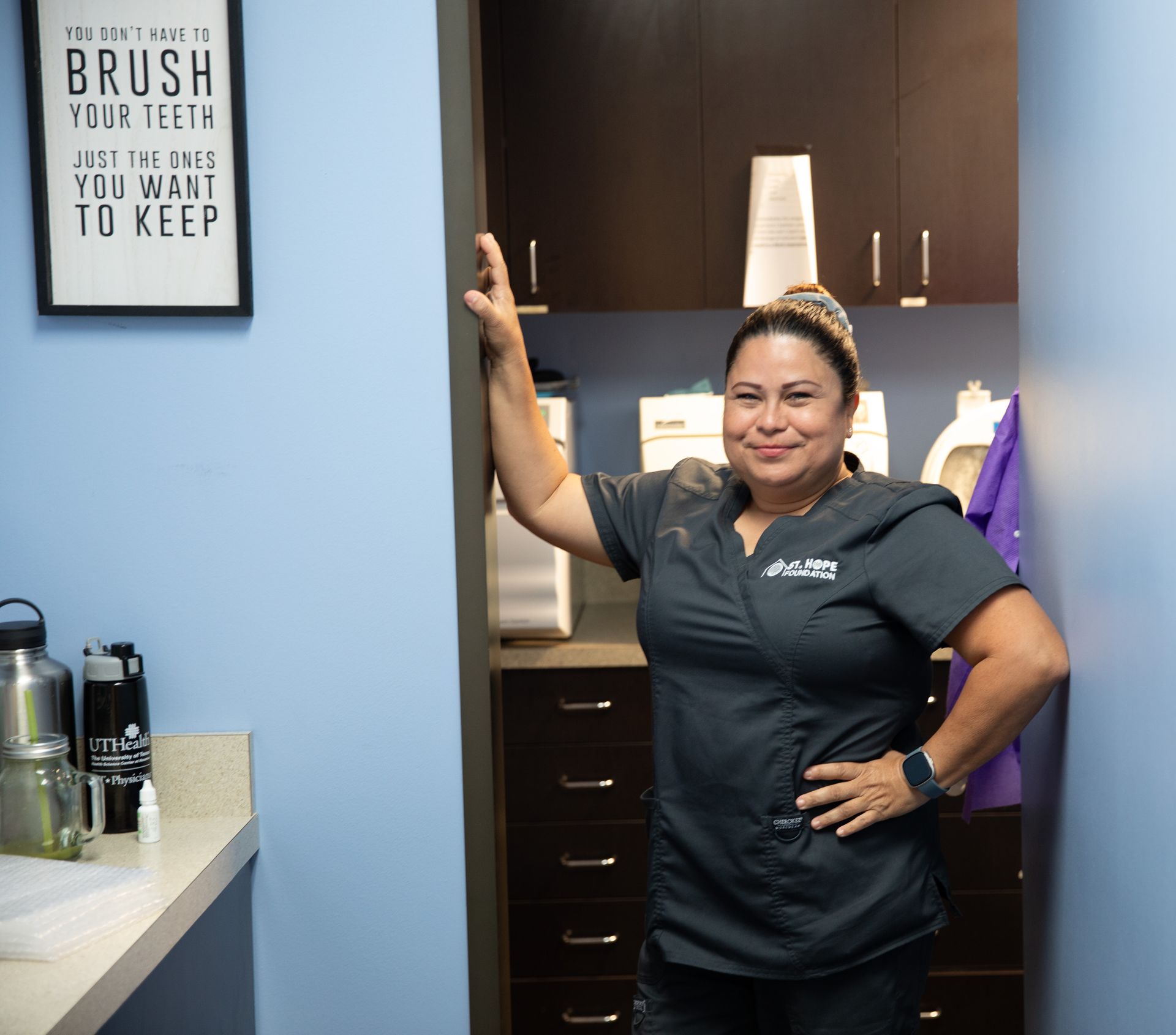 A dental professional in blue scrubs leans against a doorway in a clinic, smiling, with a motivational dental sign nearby.