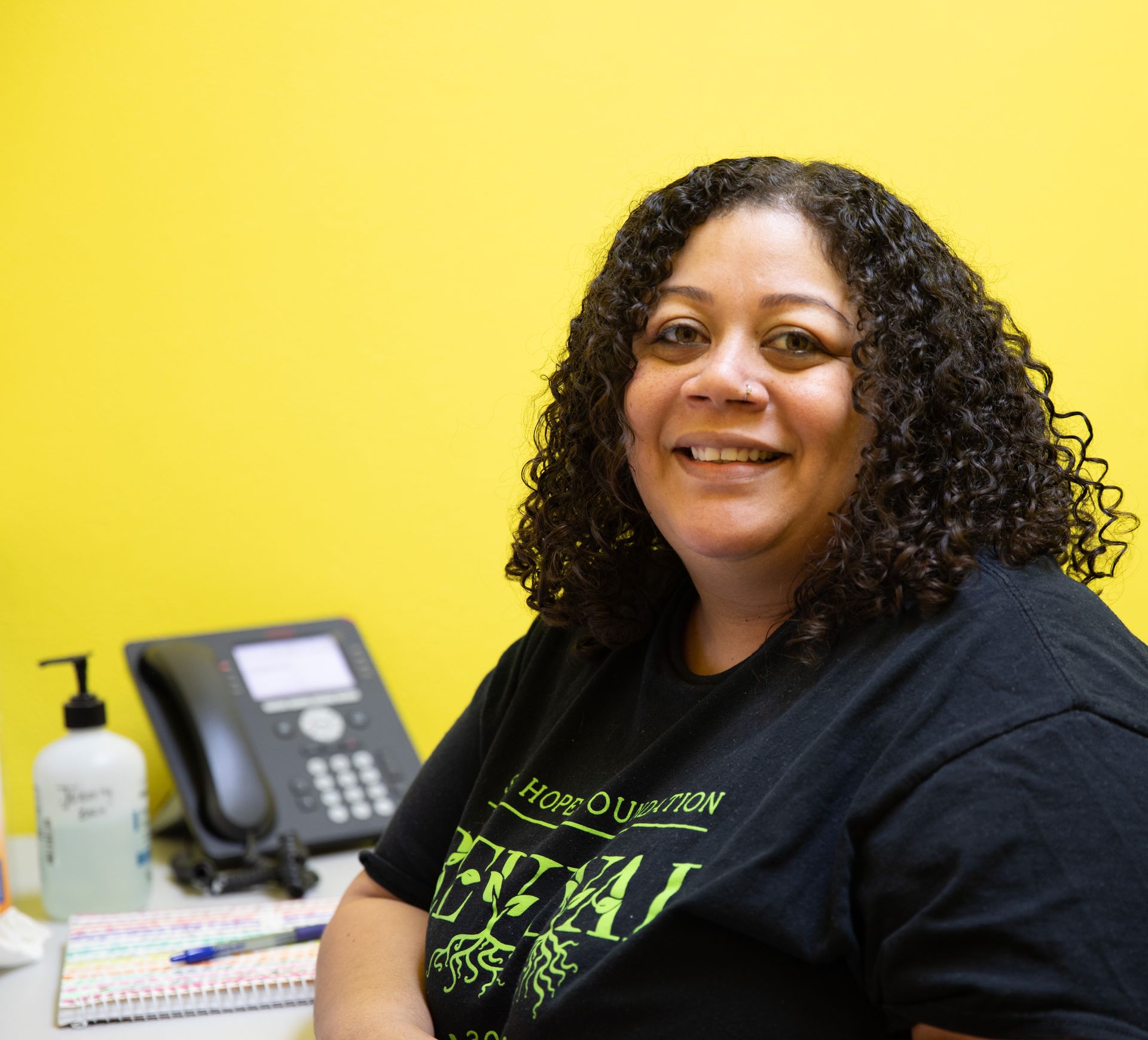 A person with curly hair smiles while sitting at a desk with a phone and sanitizer against a bright yellow wall.
