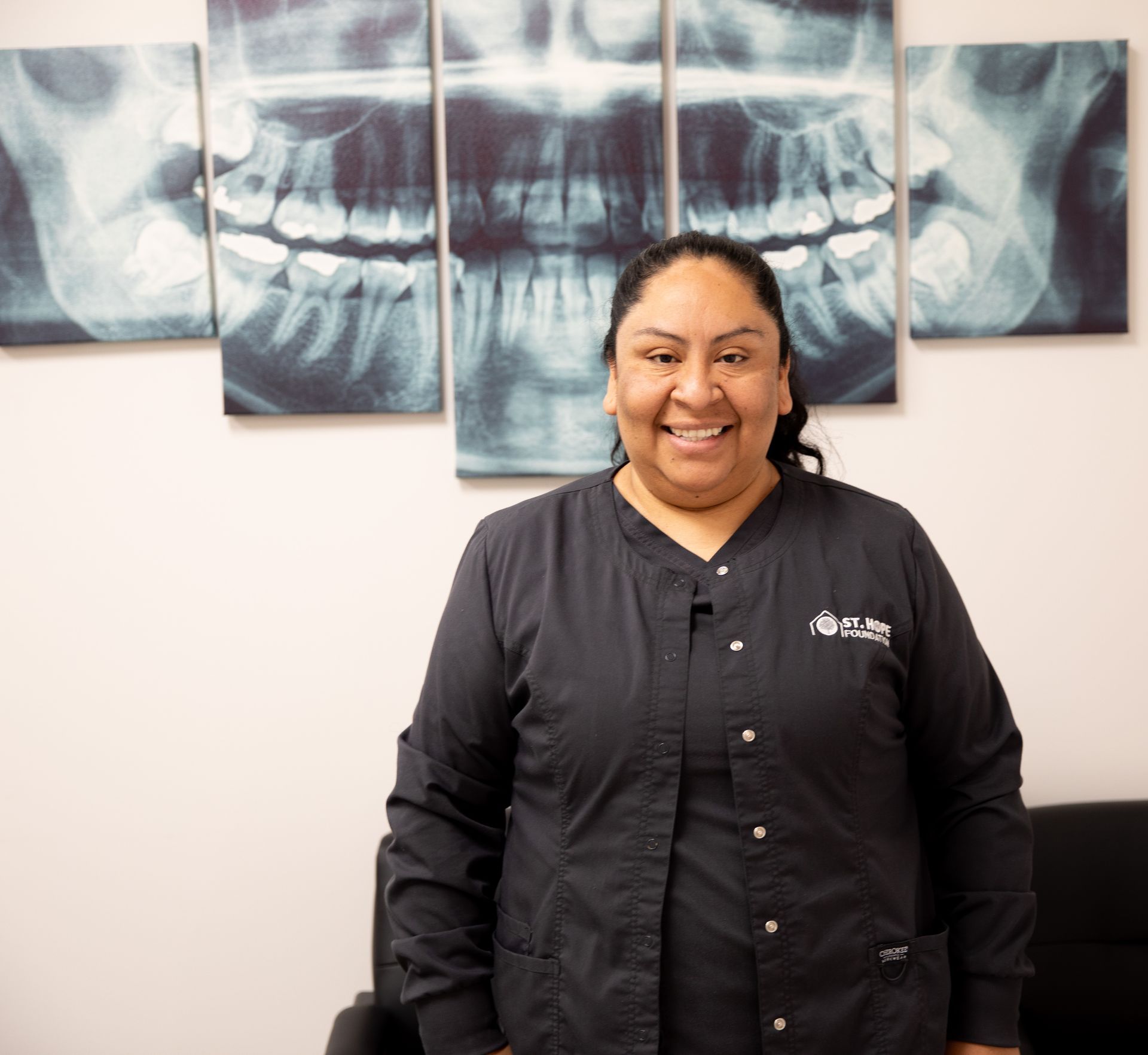 A smiling dental professional in a black scrub top stands in front of a wall decorated with a large dental X-ray print.