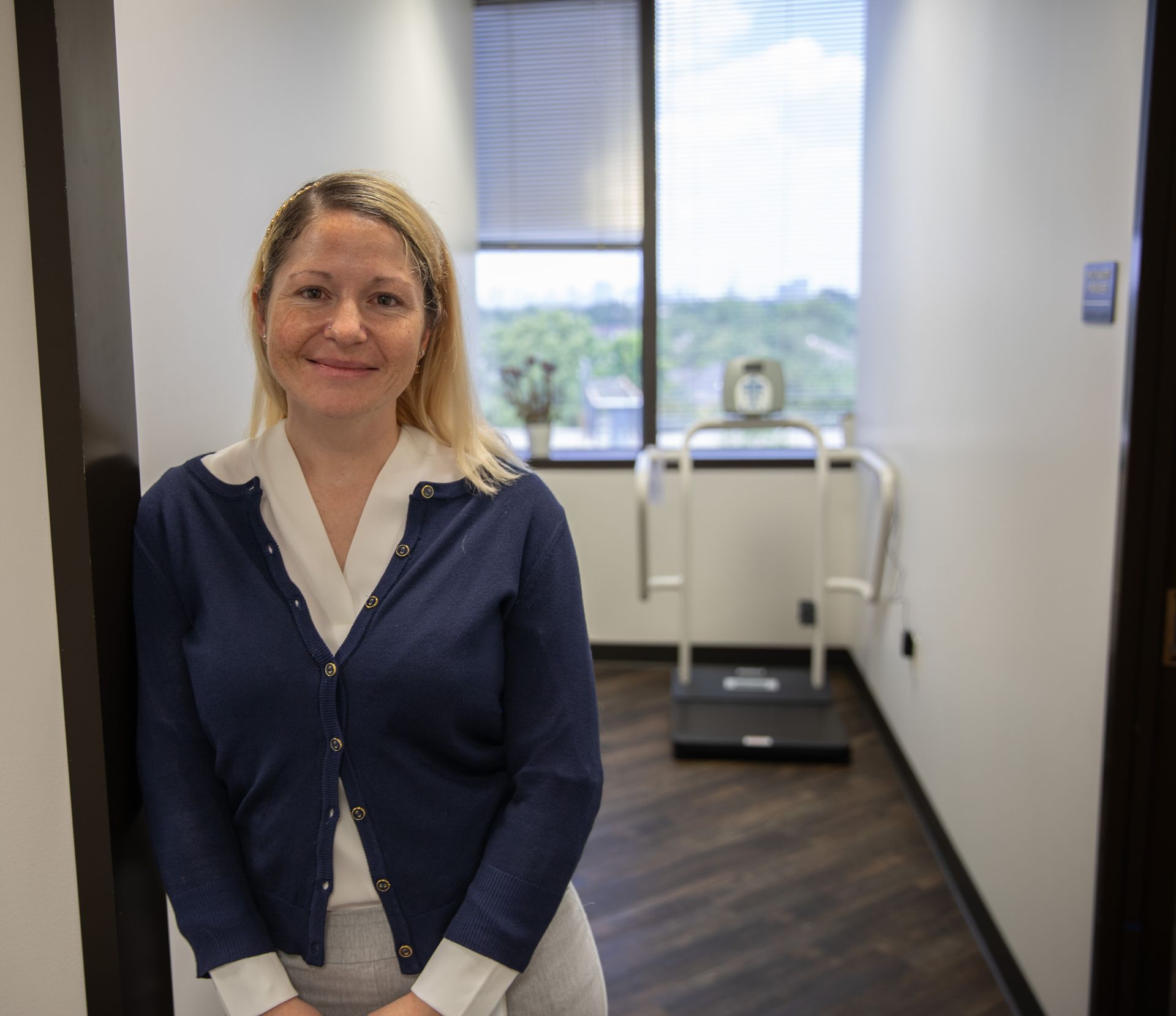 A person in a blue cardigan stands in a doorway, with a body composition scale visible in the office behind them.