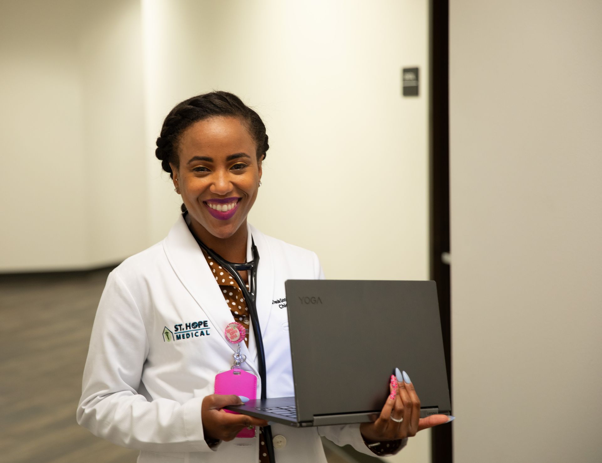 A medical professional in a white coat and stethoscope holds an open laptop in a hallway, smiling at the camera.