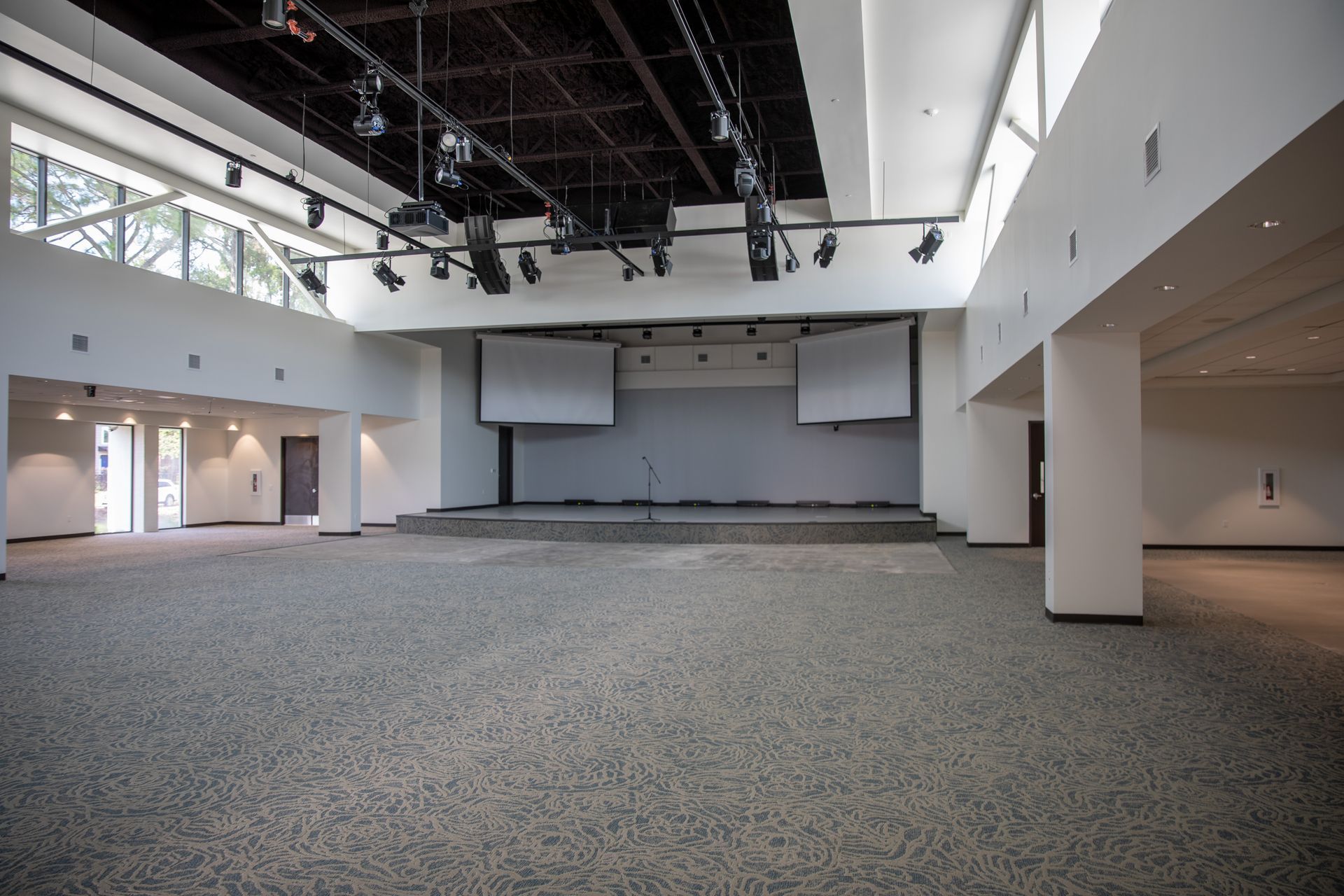 A large, empty event hall with neutral gray carpet, white walls, and a stage with two projector screens under spotlights.