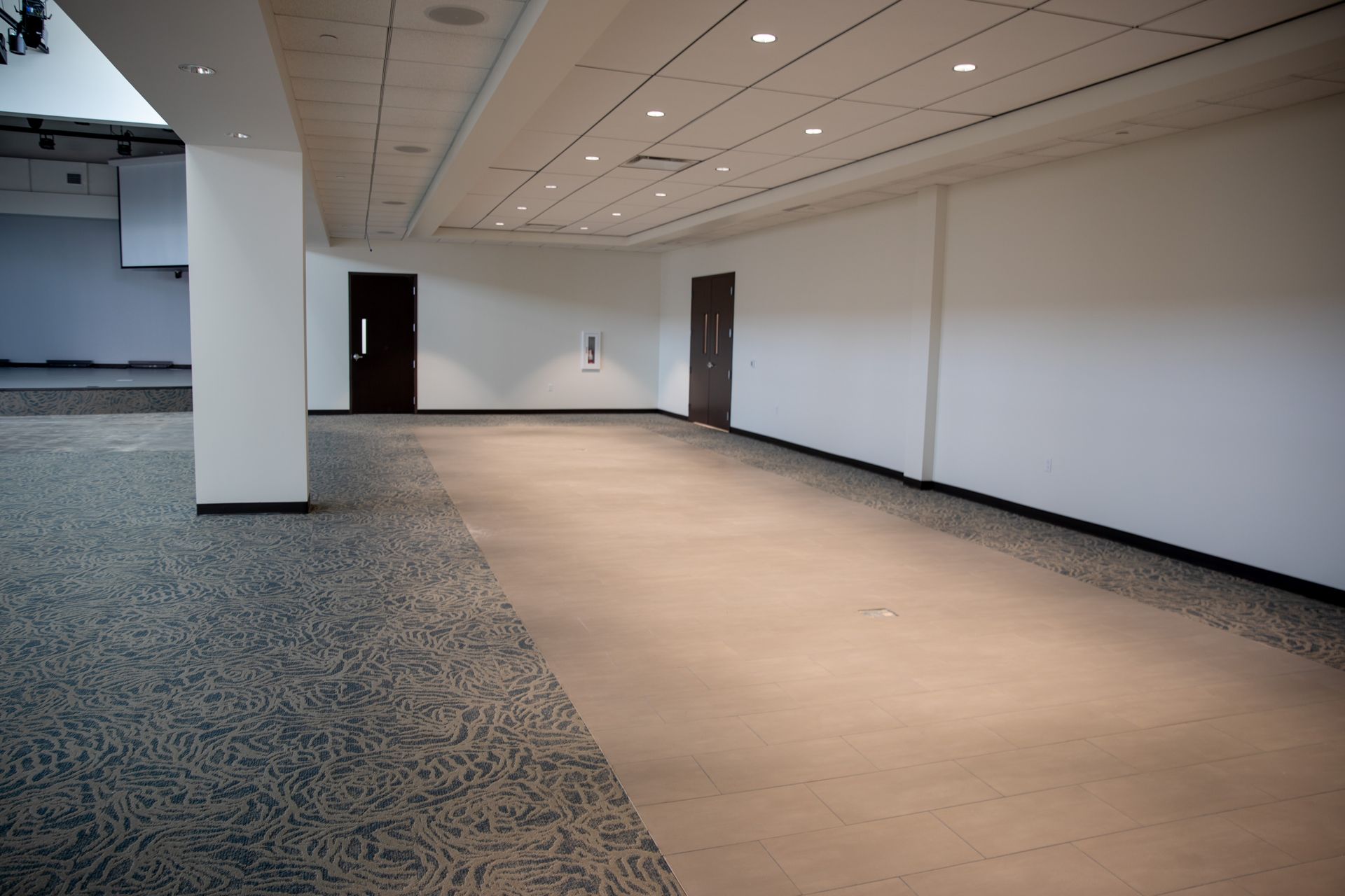 A wide, empty room features a combination of patterned carpet and smooth light flooring with recessed ceiling lights.