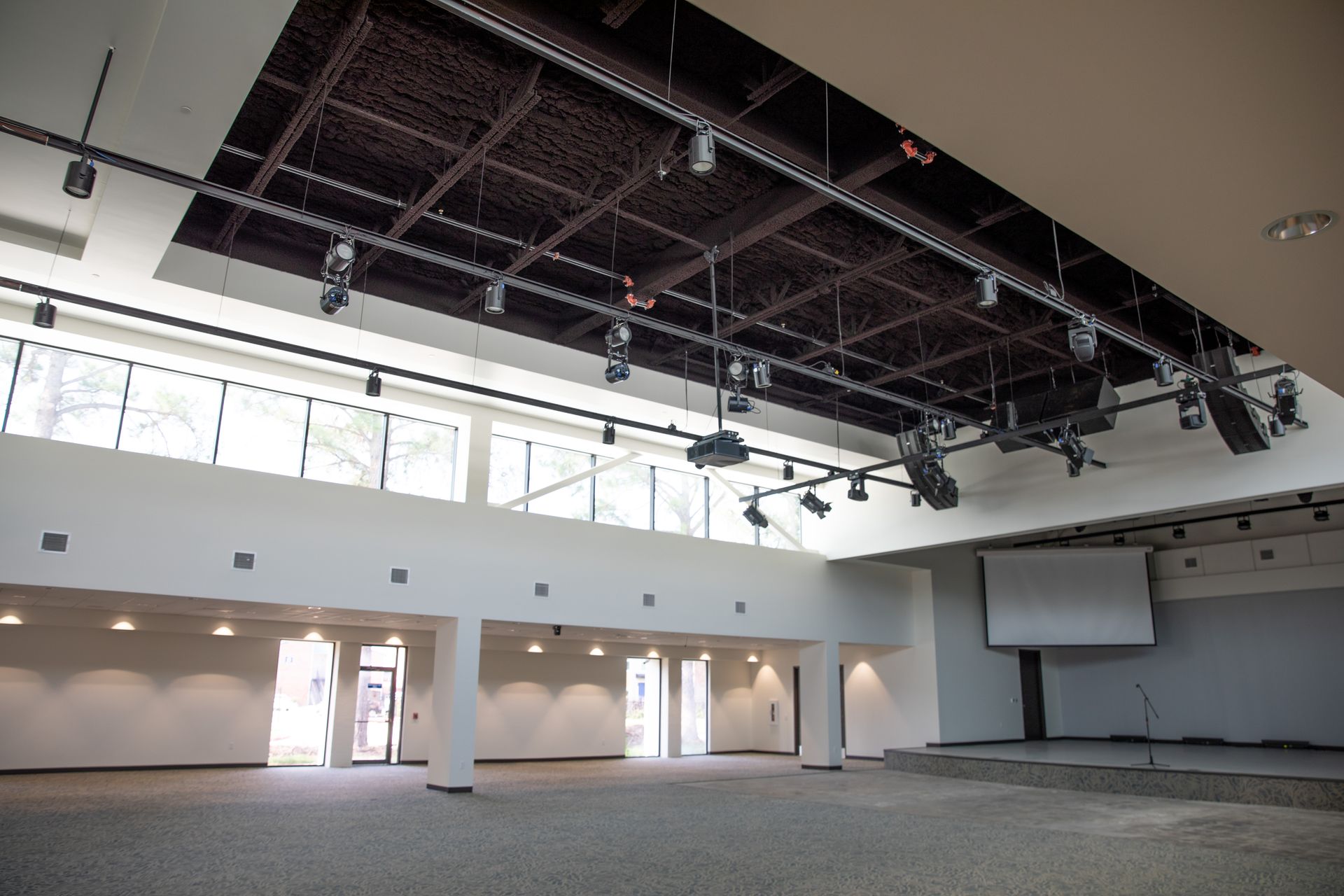A spacious, modern event hall with a high, dark wooden ceiling, stage, projector screen, and soft overhead lighting.