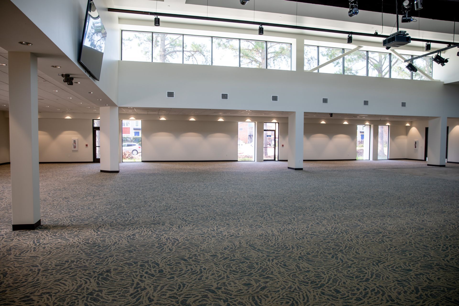 A spacious, empty event hall with high ceilings, large windows, pillars, and gray patterned carpeting.