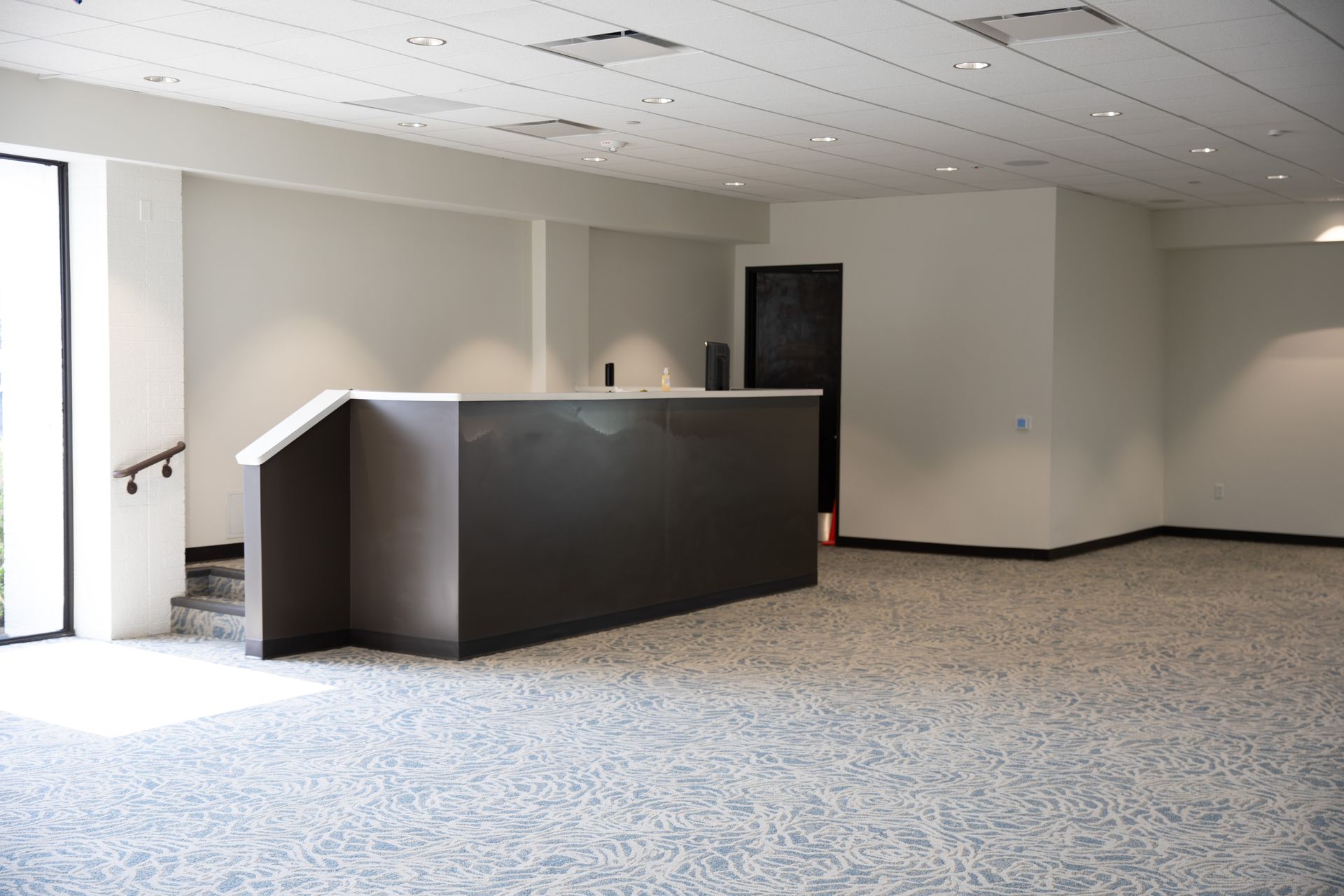 A large, empty lobby with a long, dark-grey reception desk, patterned grey carpet, white walls, and recessed lighting.
