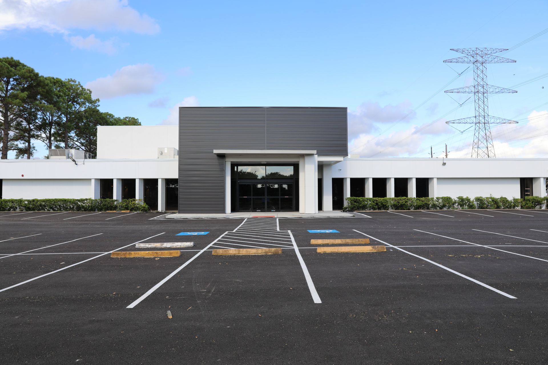 Single-story white commercial building with a black paneled entryway and a large, empty asphalt parking lot.
