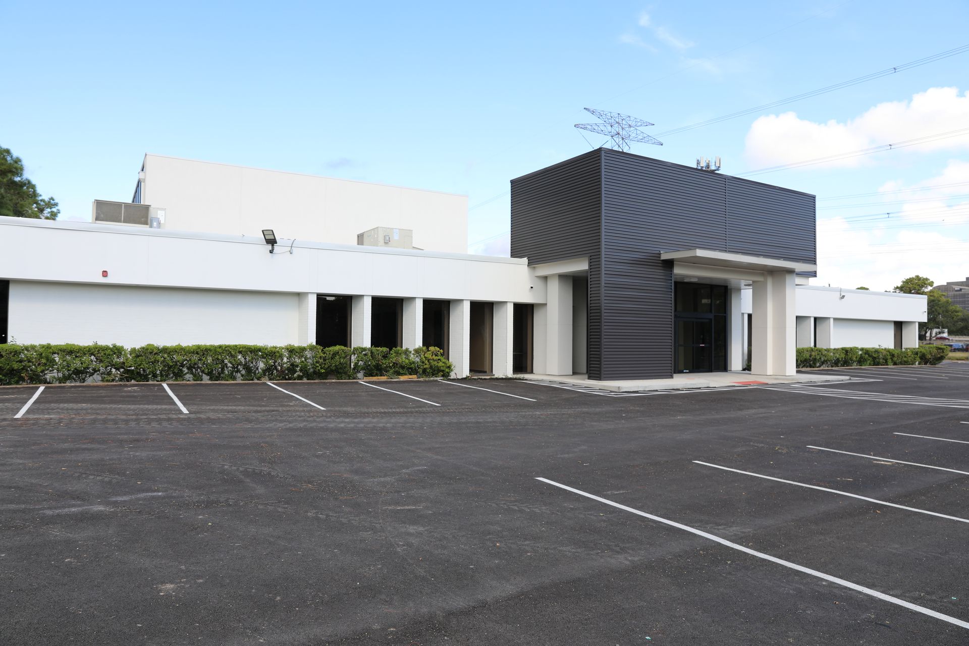 A wide, low-rise building with white walls and a prominent black, angular entrance structure, fronted by a large parking lot.