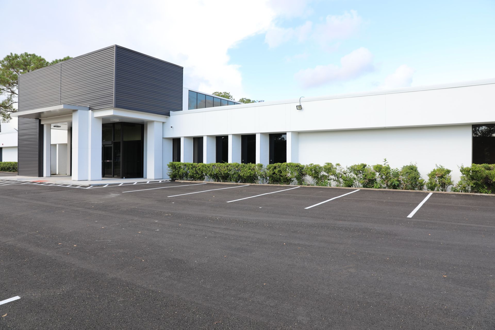 A single-story office building with a dark gray entrance, white facade, and landscaped bushes next to a paved parking lot.