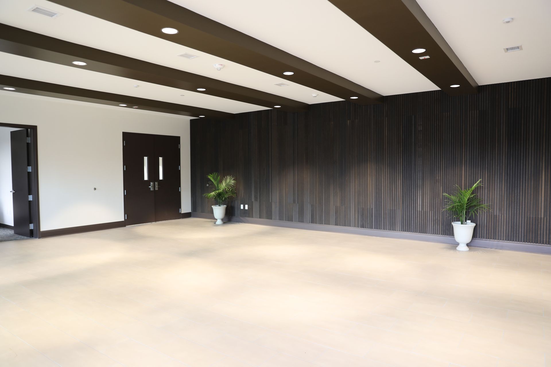 An empty event hall with light carpet, dark wooden wall accents, brown double doors, and two potted plants.