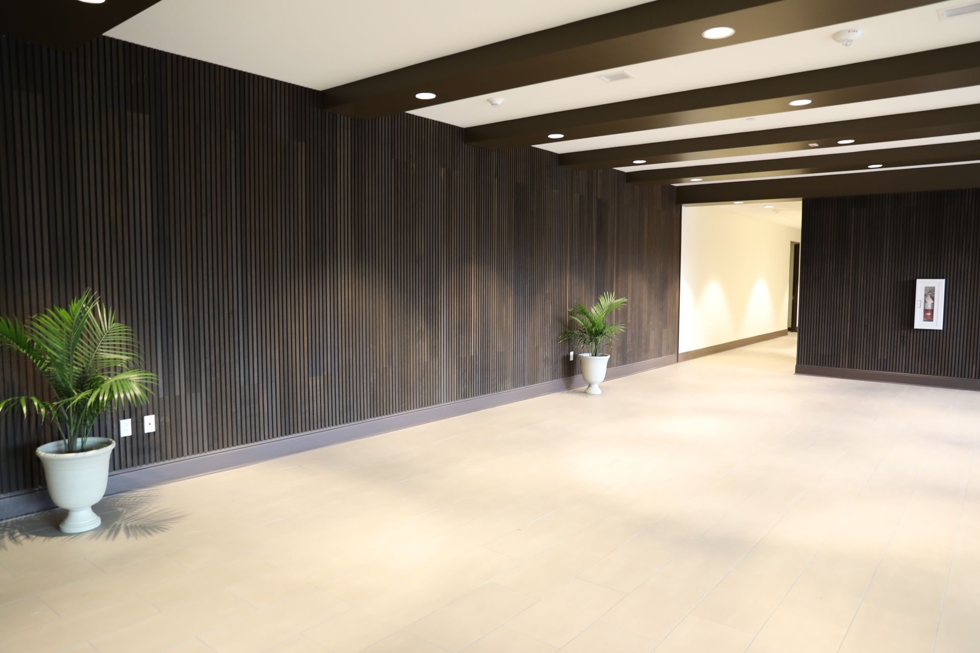 A wide, modern hallway with light-colored flooring, dark vertical wall slats, recessed lighting, and two potted plants.