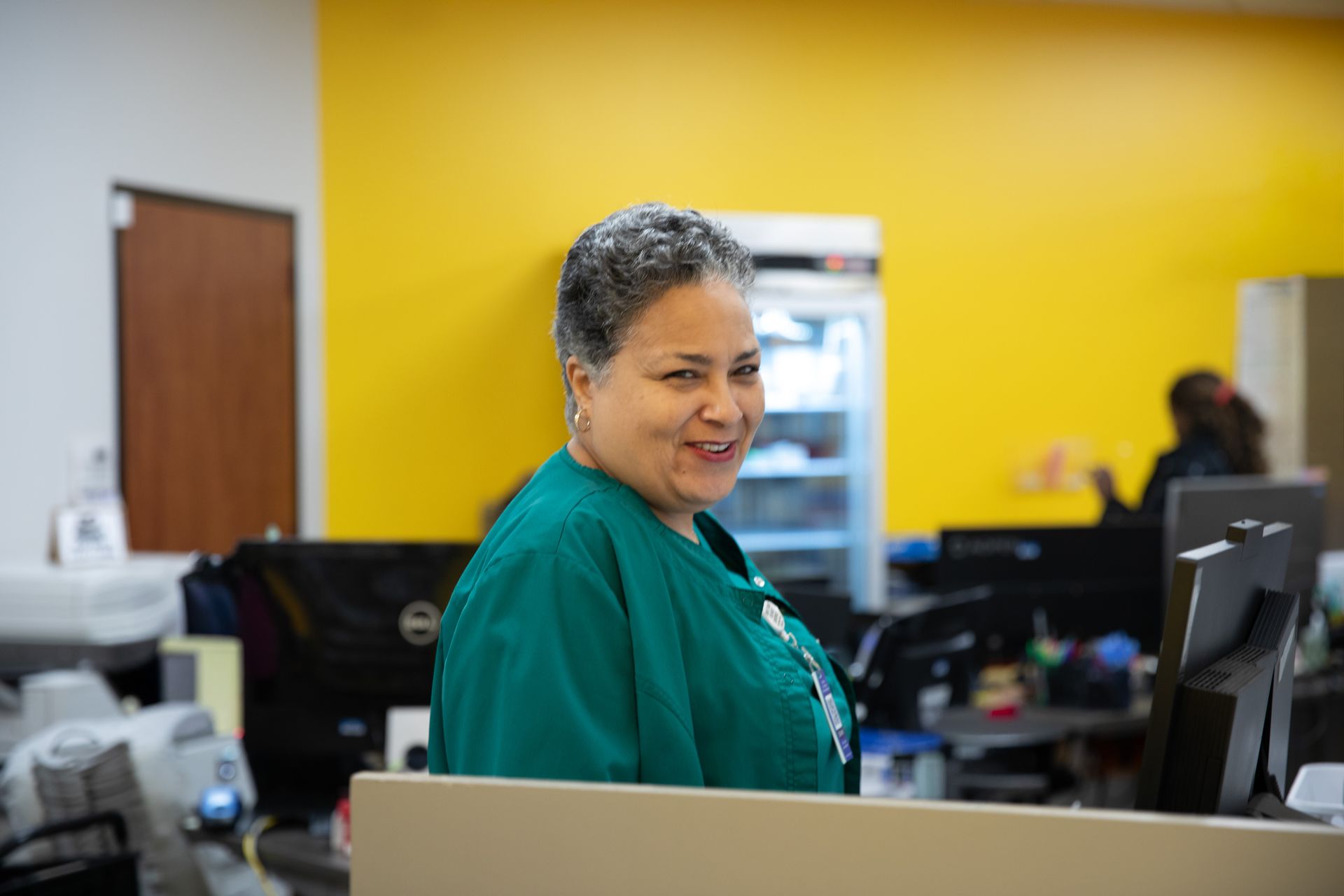 A person in a green uniform smiles at the camera from behind a desk in an office with a bright yellow wall.