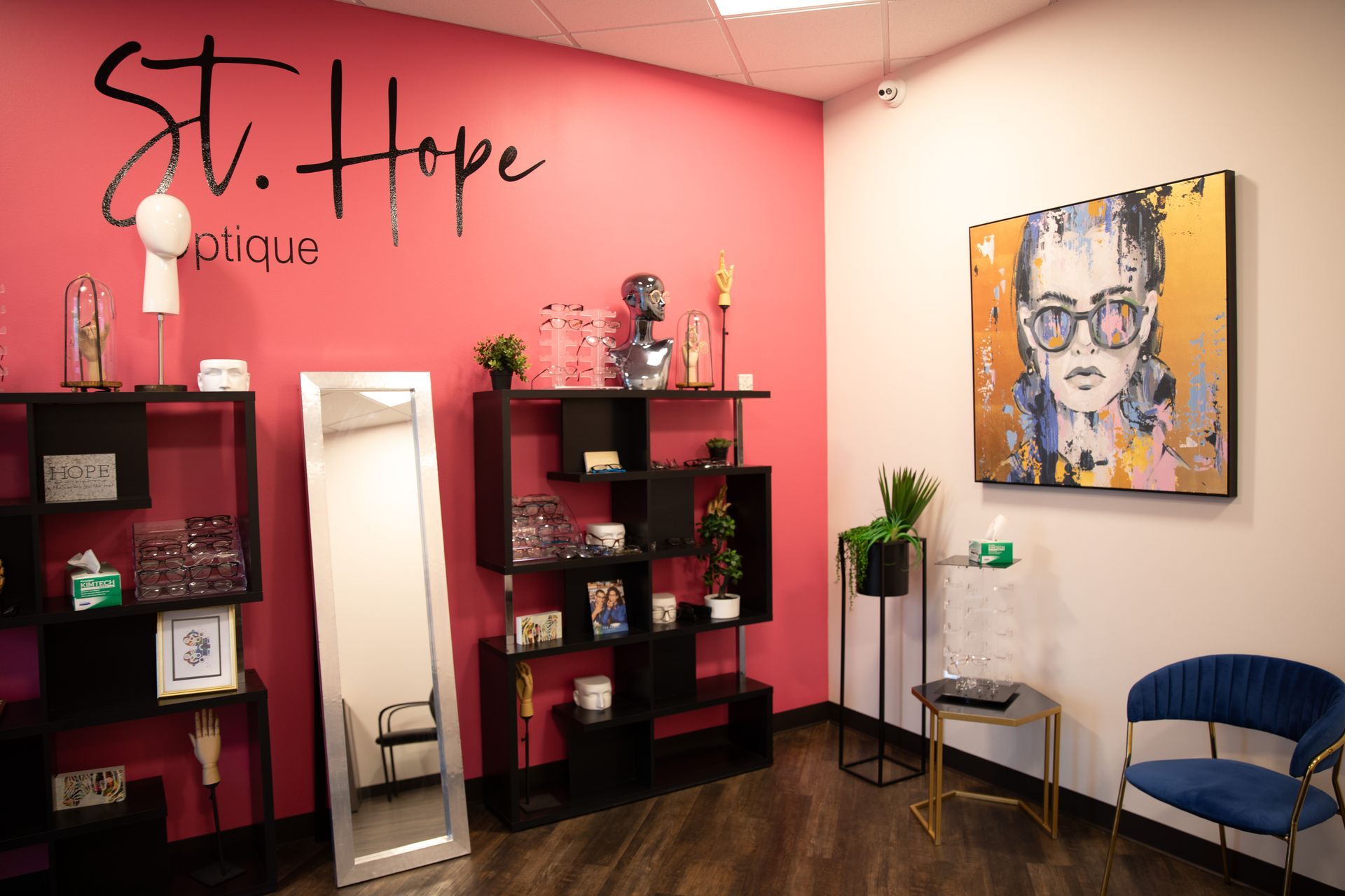 St. Hope Optique boutique with black shelving, a floor mirror, decorative items, and portrait art on a pink wall.