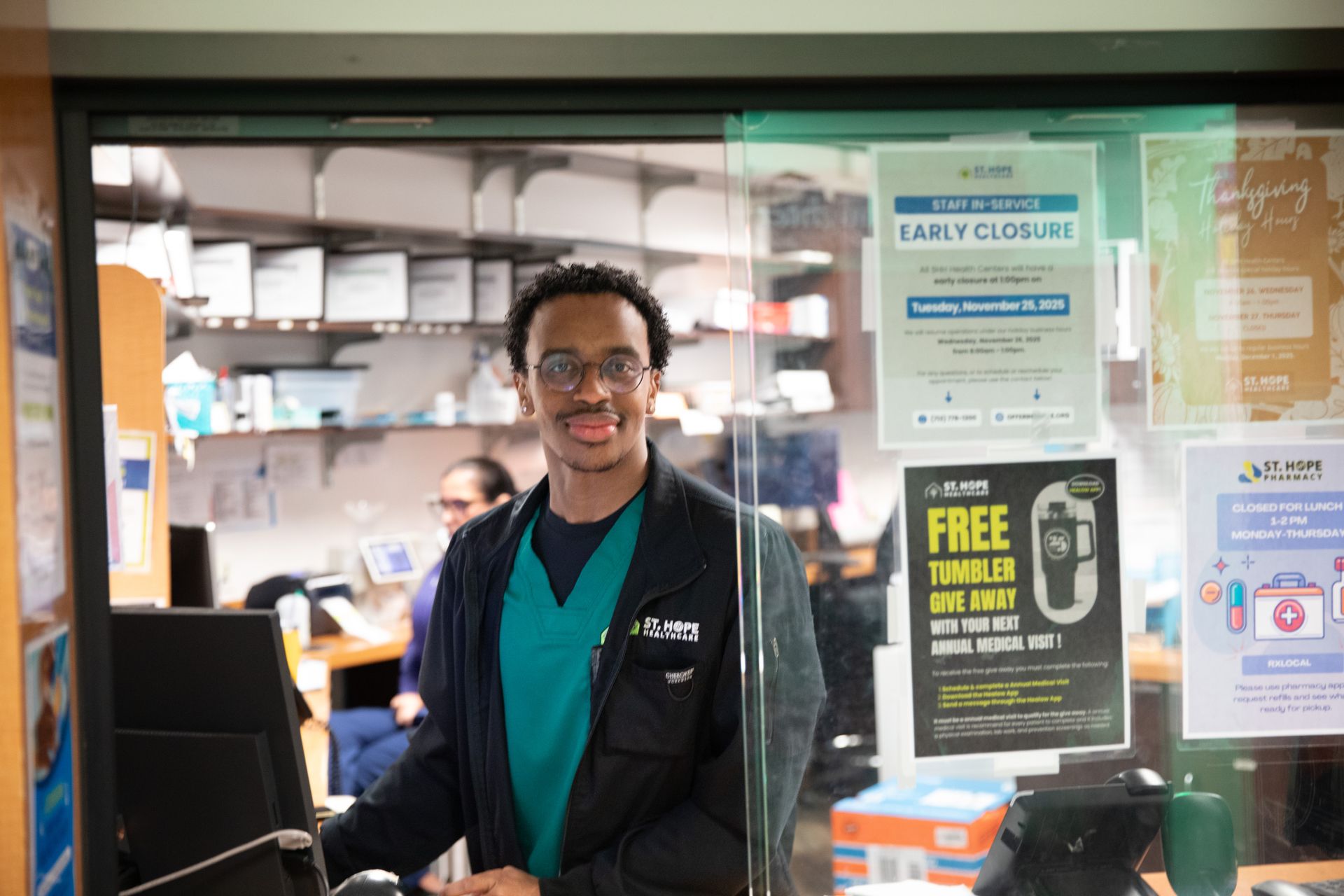 A staff member in a teal scrub top and black jacket smiles while working behind a glass pharmacy counter.