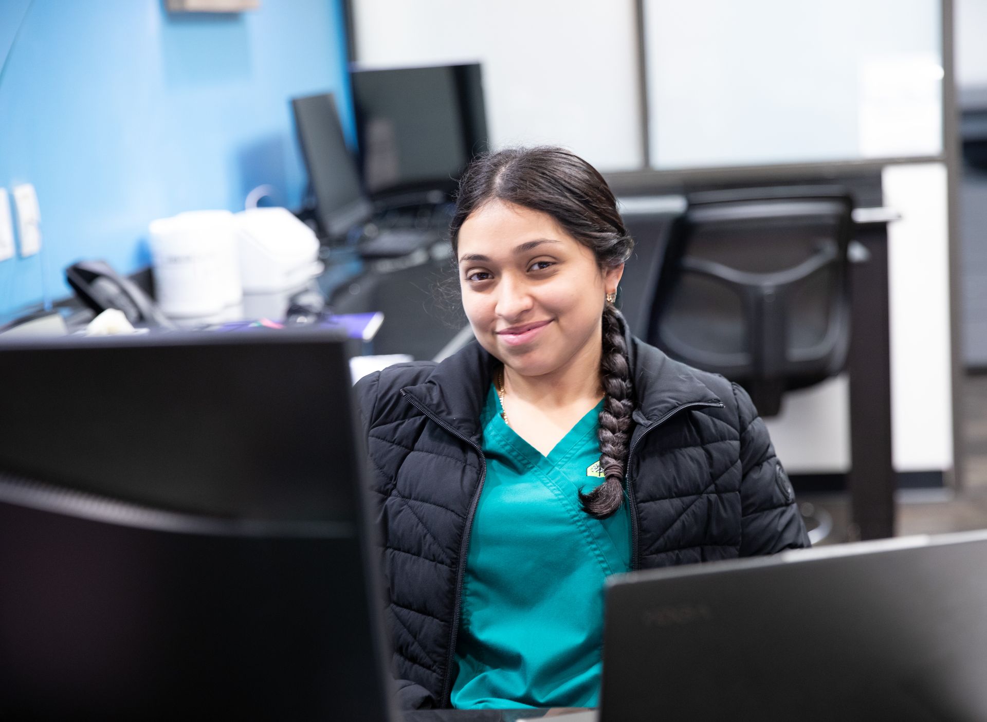 A person wearing a teal scrub top and black jacket sitting at an office desk with computers, smiling at the camera.