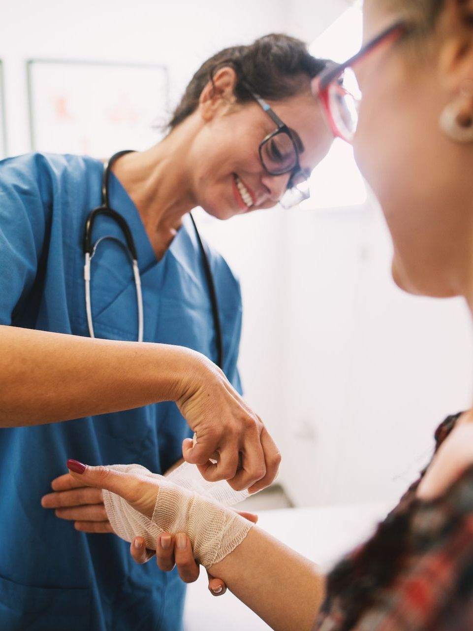 A medical professional in blue scrubs applies a bandage to a patient's wrist in a clinical setting.