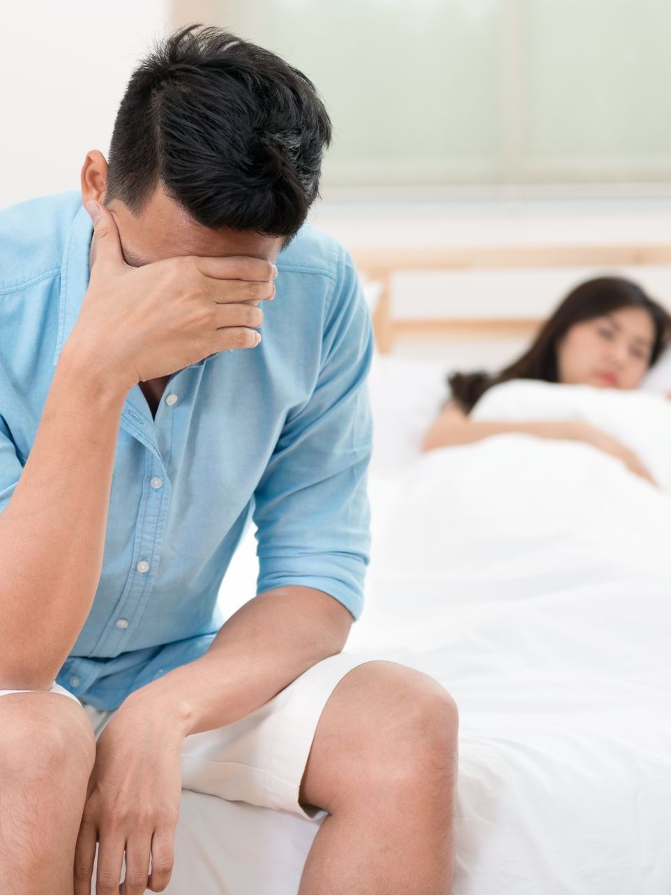 A distressed person sits on a bed with their hand on their forehead, while another person sleeps in the background.