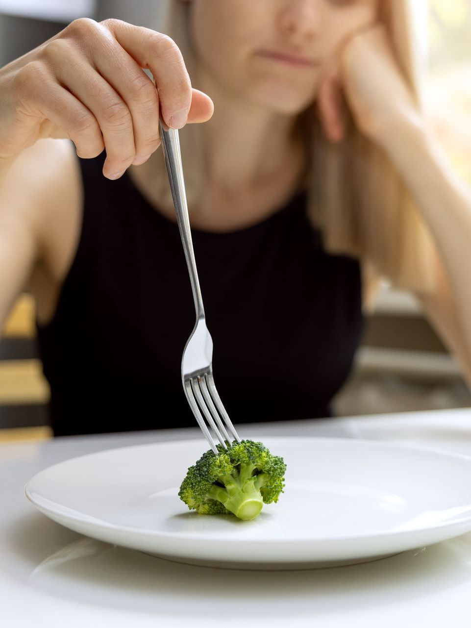 A person sits at a table, looking unenthusiastic while holding a fork with a single piece of broccoli on a white plate.