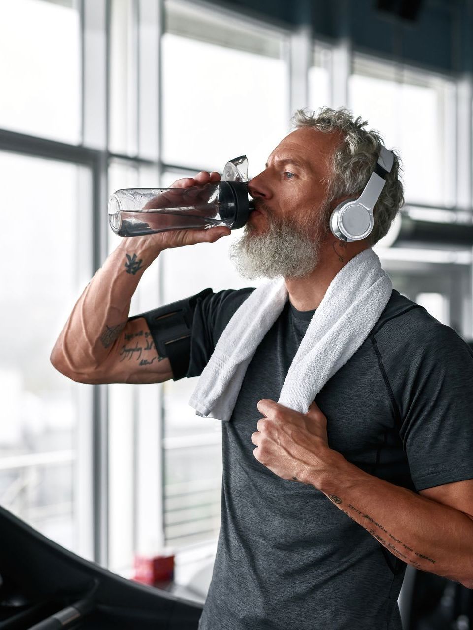 A person with a beard and headphones drinks from a water bottle in a gym, wearing a gray shirt and a white towel.