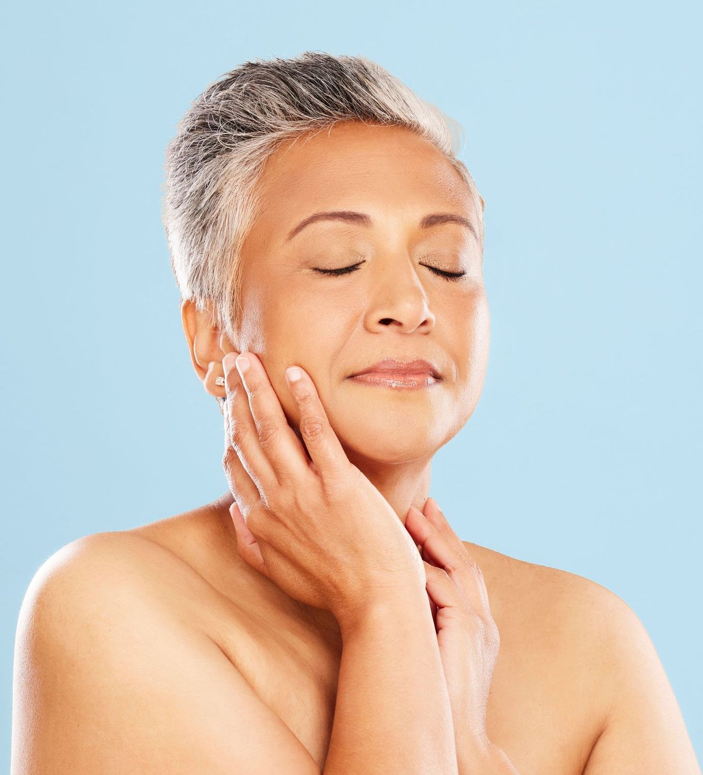 A person with short grey hair, eyes closed, gently touching their face and neck against a light blue background.