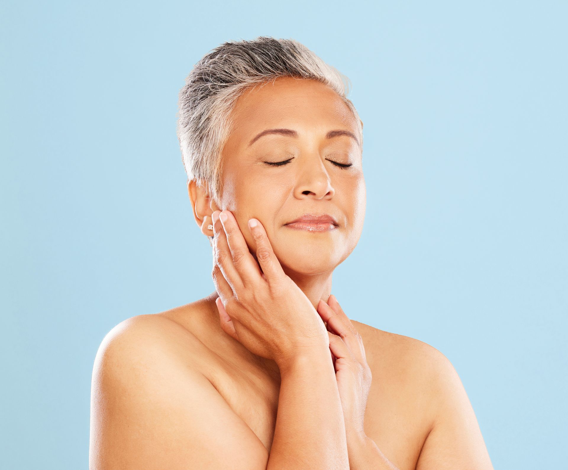A person with short grey hair, eyes closed, gently touching their face against a light blue background.