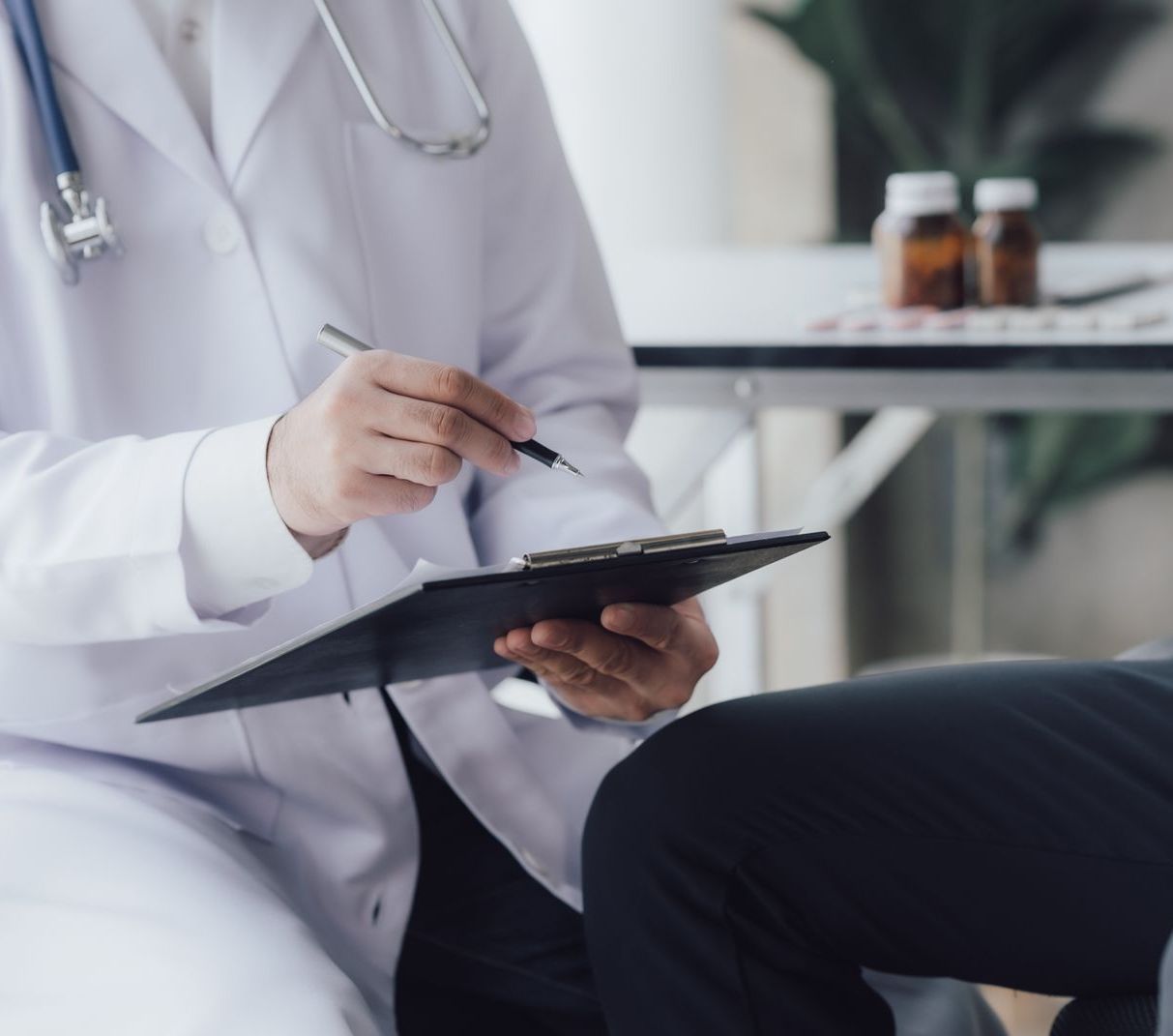 A doctor in a white coat writes on a clipboard during a consultation in an office setting.