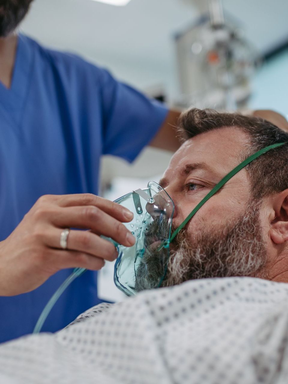 A medical professional in blue scrubs places an oxygen mask over the face of a patient wearing a patterned hospital gown.