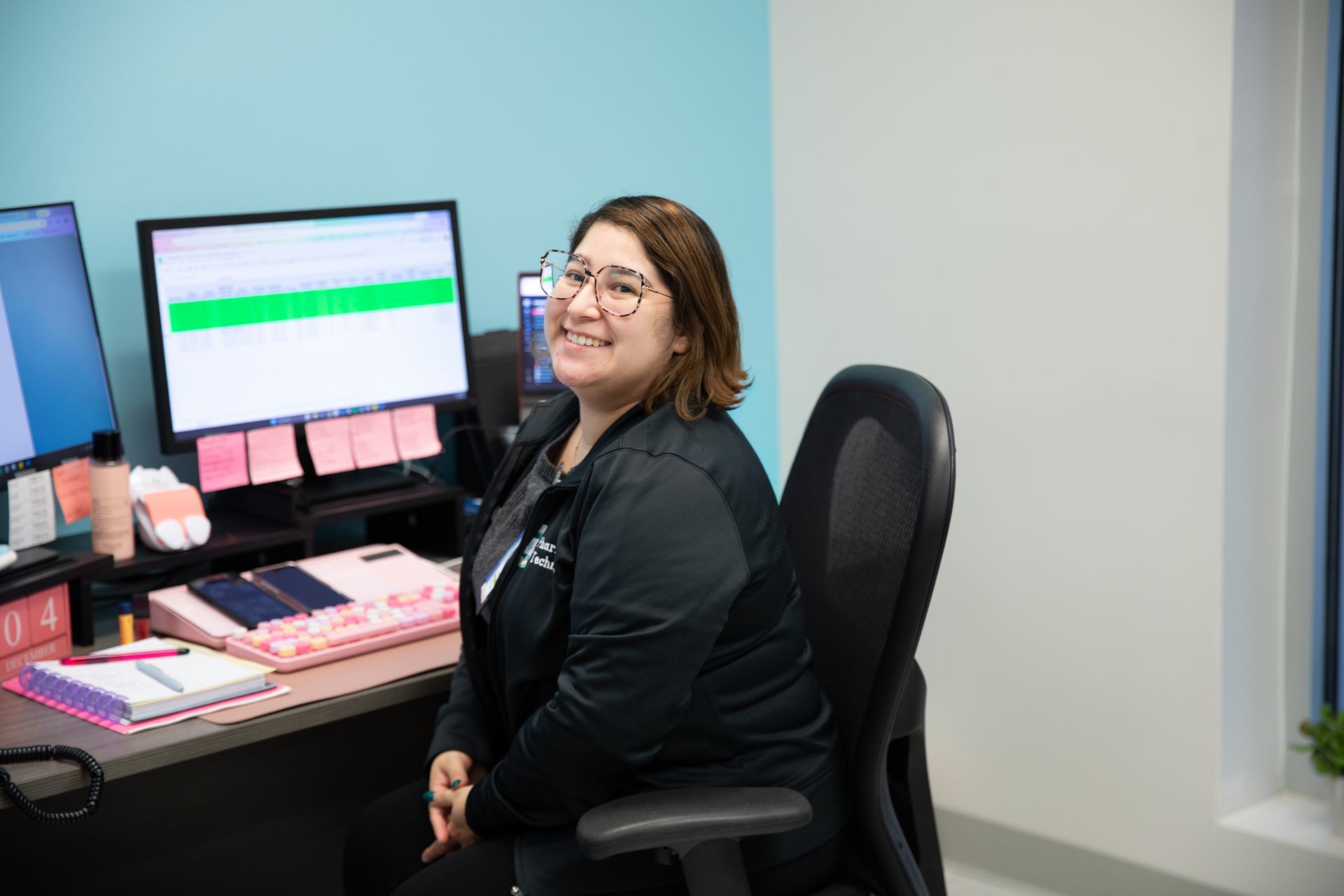 A smiling professional wearing glasses sits in an office chair at a desk with two computer monitors and a notepad.