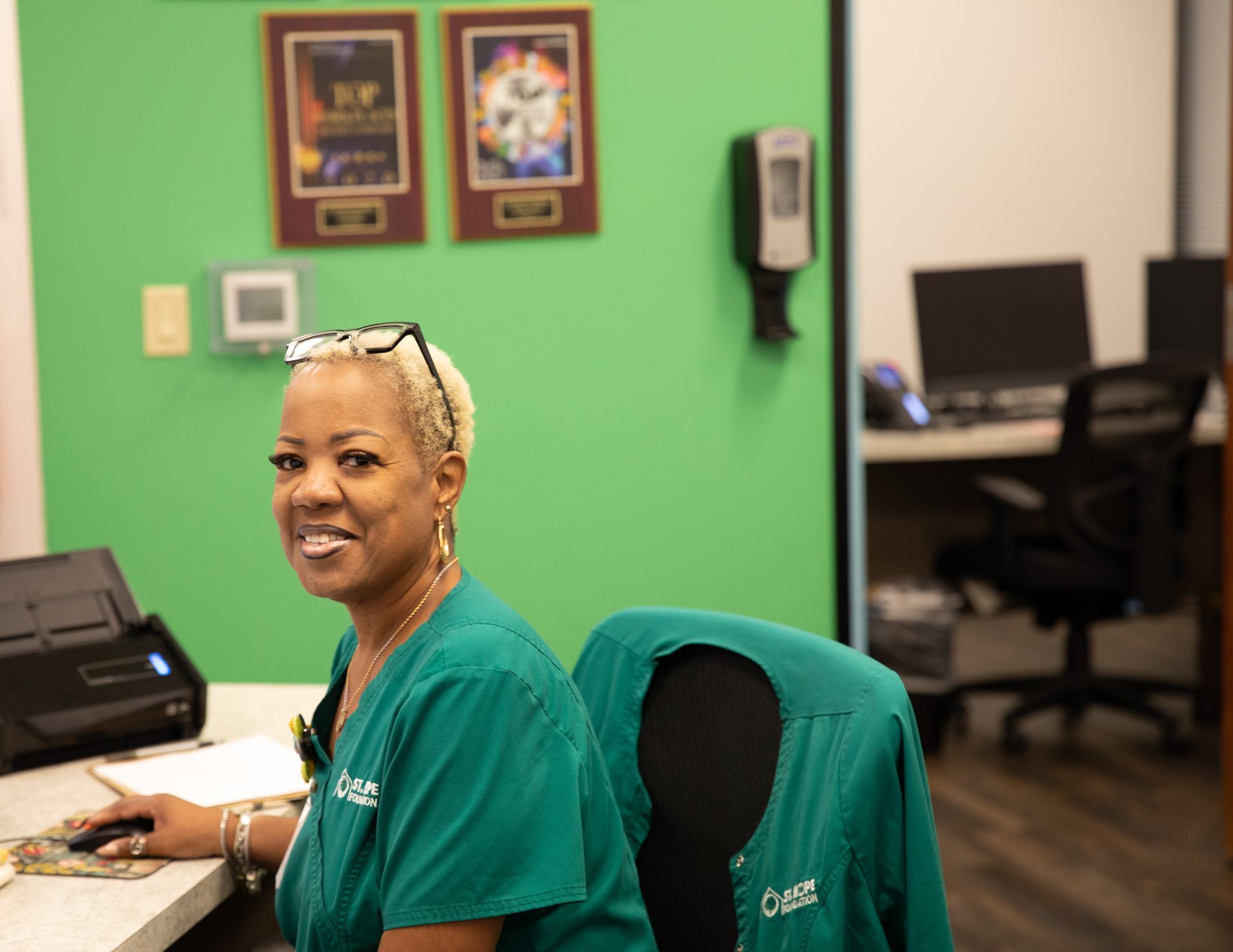 A person in a green uniform sits at a desk, working at a computer in an office with bright green walls and wall decor.