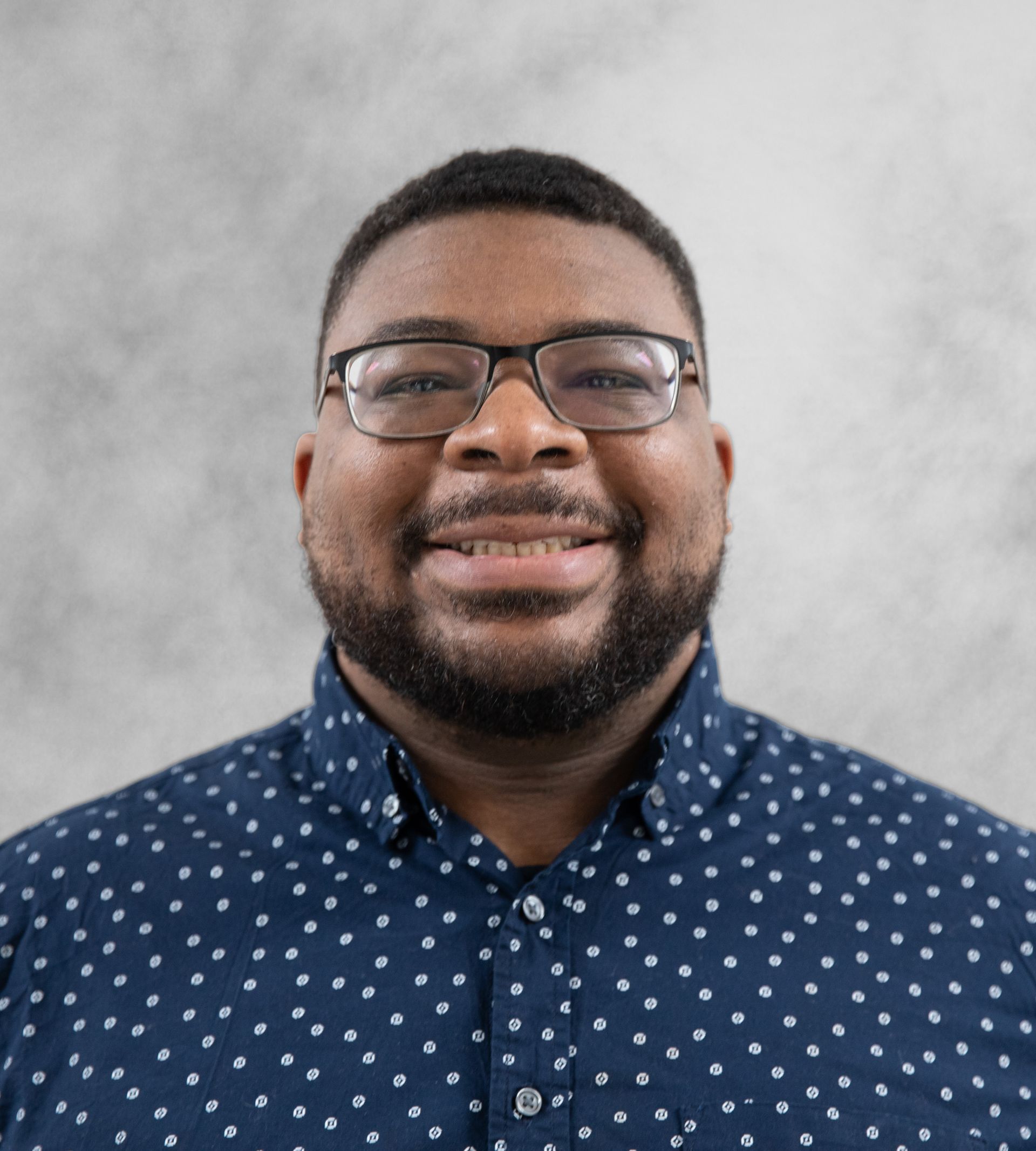 A person wearing glasses and a blue button-down shirt with a white dotted pattern, smiling against a gray background.