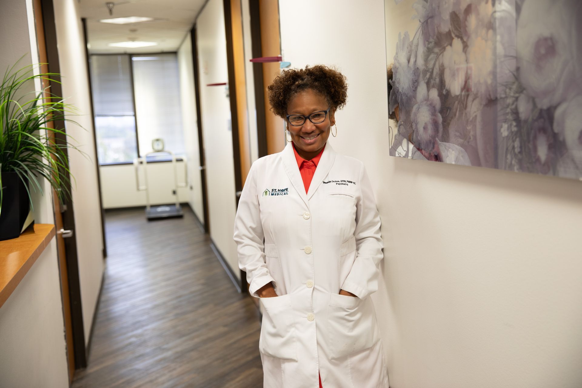 A professional in a white lab coat standing in a hallway with hands in pockets, smiling at the camera.
