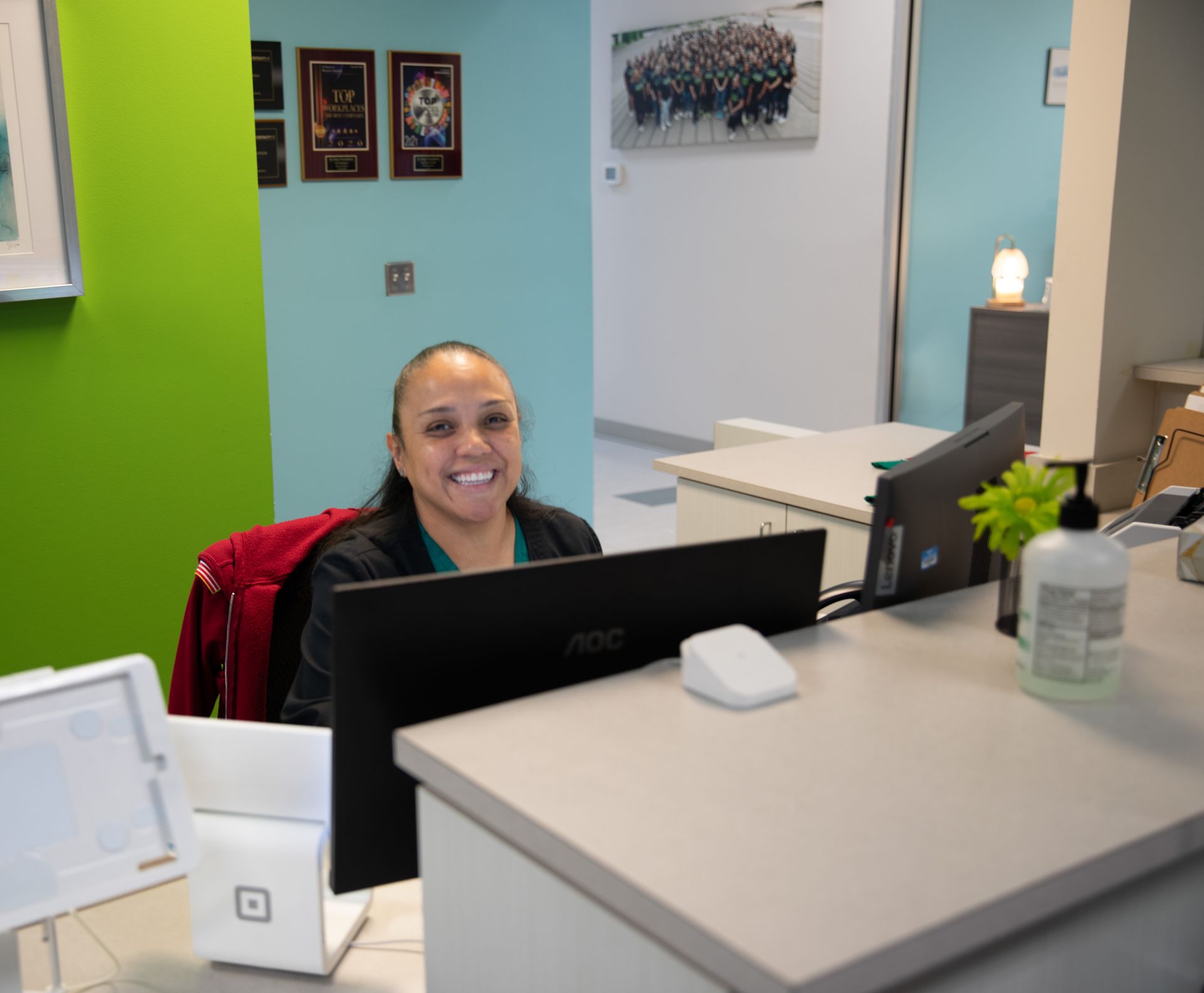 A person smiling behind an office reception desk with a computer monitor, desk accessories, and colored walls.