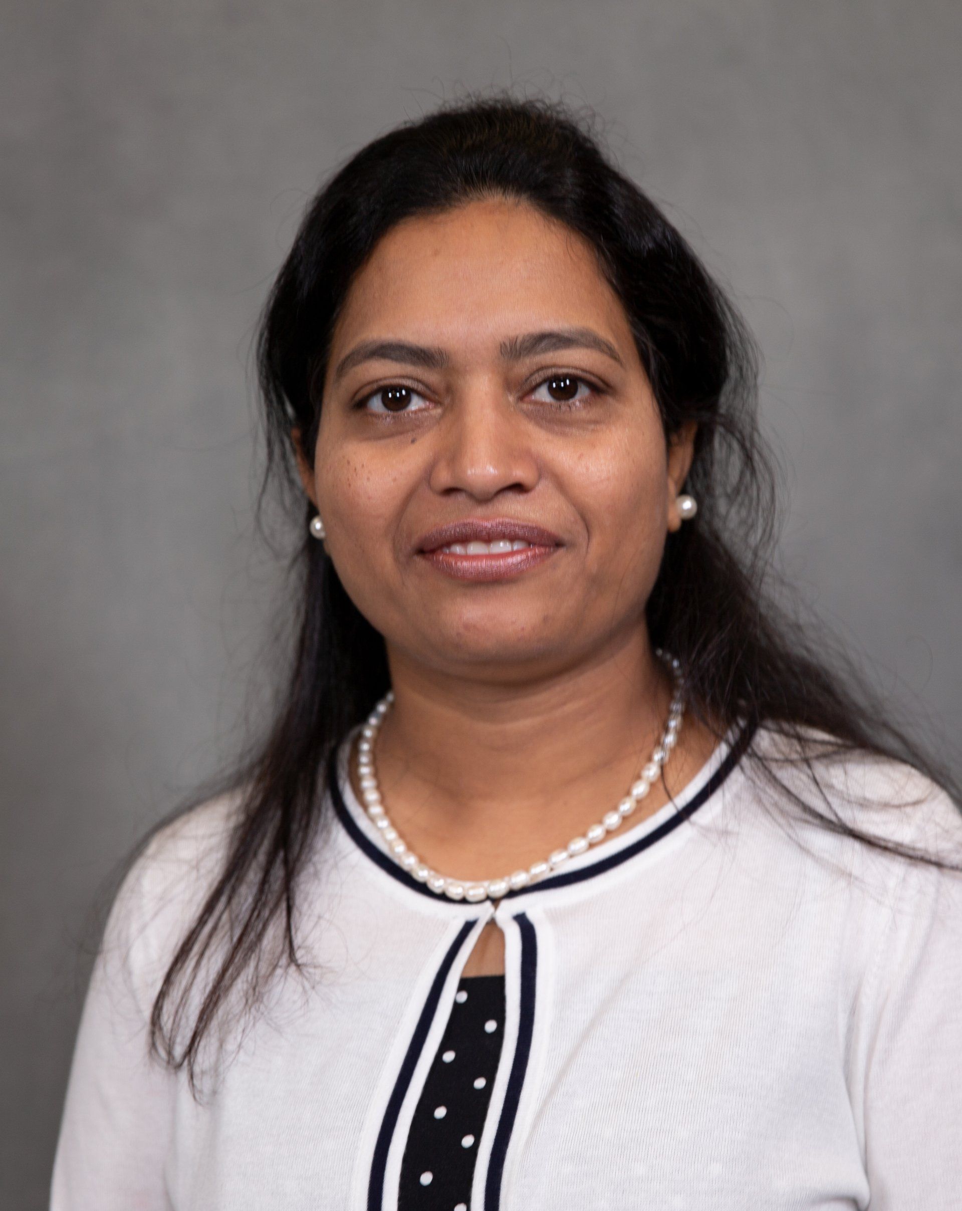 Headshot of a person with long dark hair, wearing a white cardigan over a polka-dot top and a pearl necklace.