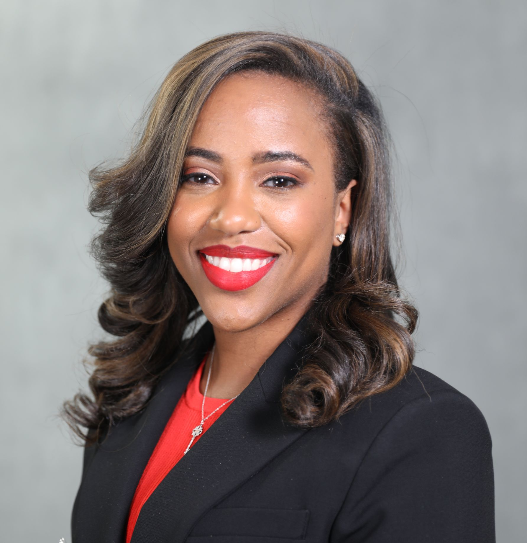 A smiling woman with long brown hair, wearing a black blazer over a red top, against a gray background.