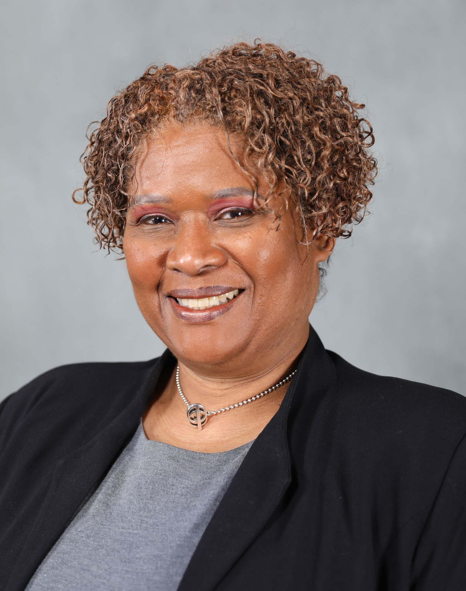 A smiling person with curly hair, wearing a gray shirt, a dark blazer, and a beaded necklace against a gray background.