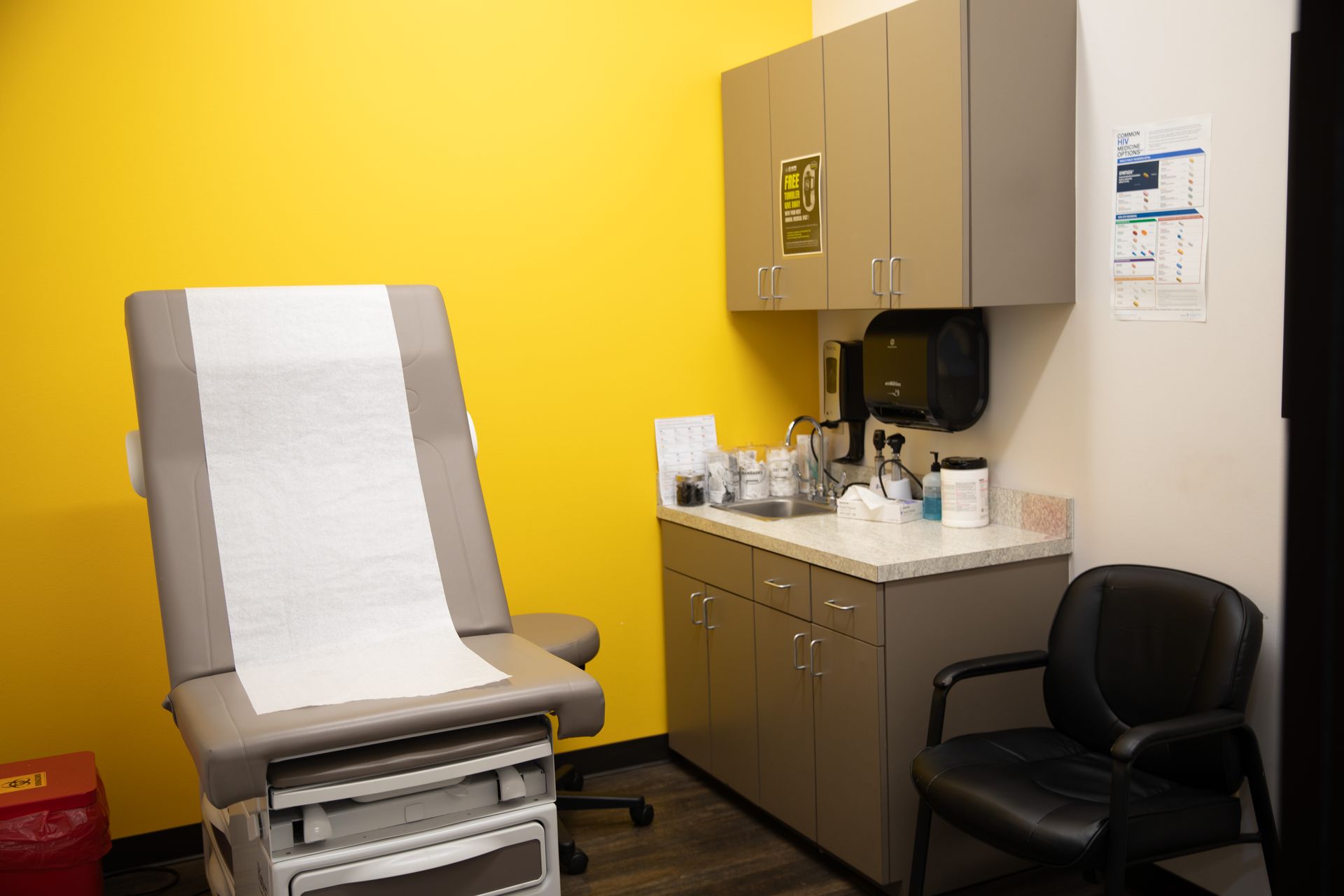 An exam room with a bright yellow wall, a gray medical exam table, a counter with supplies, and a black office chair.