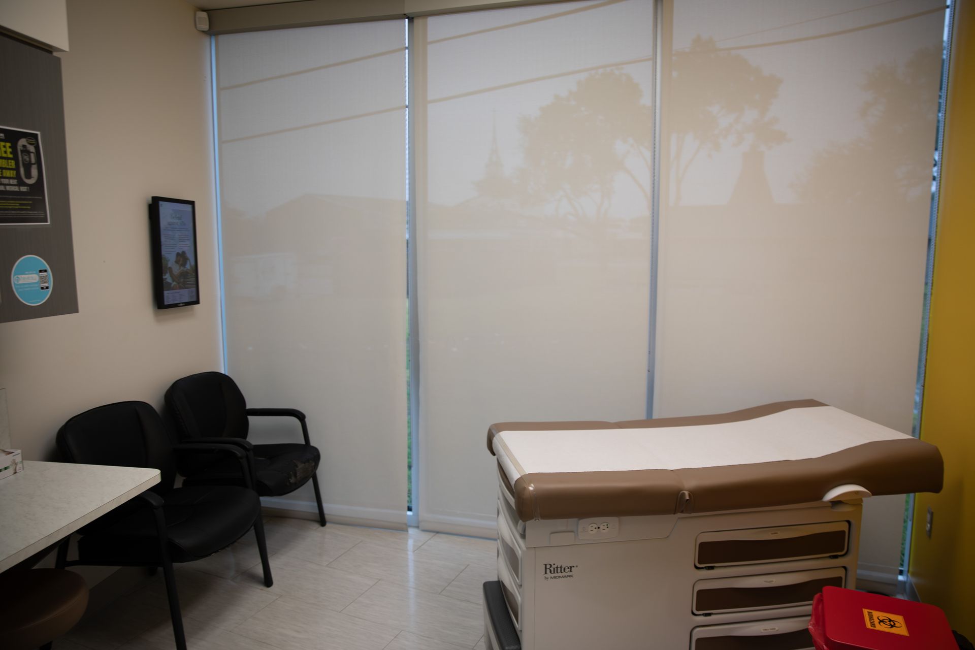 A medical examination room with an exam table, two black chairs, a side desk, and floor-to-ceiling windows with shades.