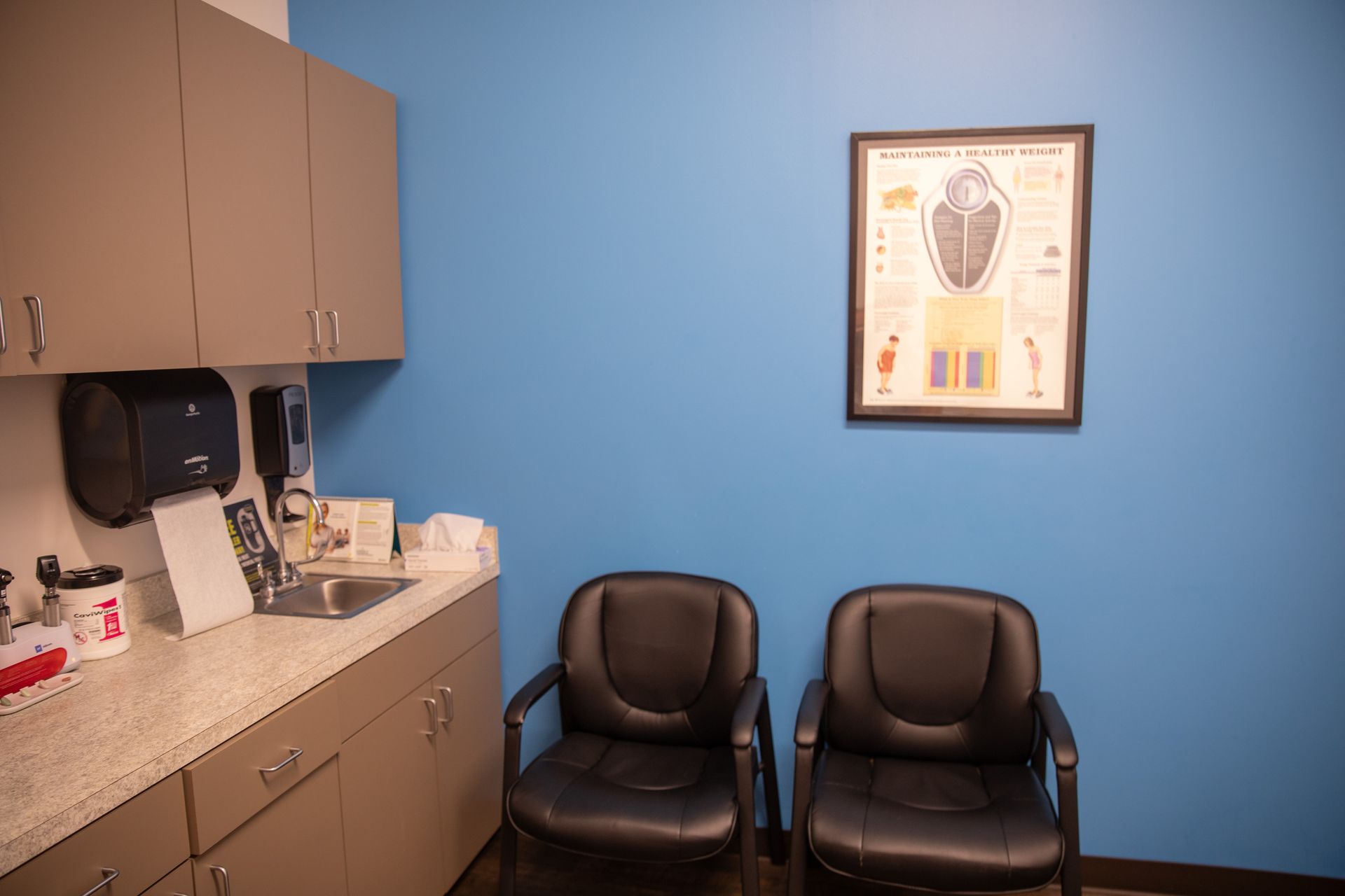 A medical examination room with tan cabinets, a sink, two black chairs, and a framed human anatomy chart on a blue wall.
