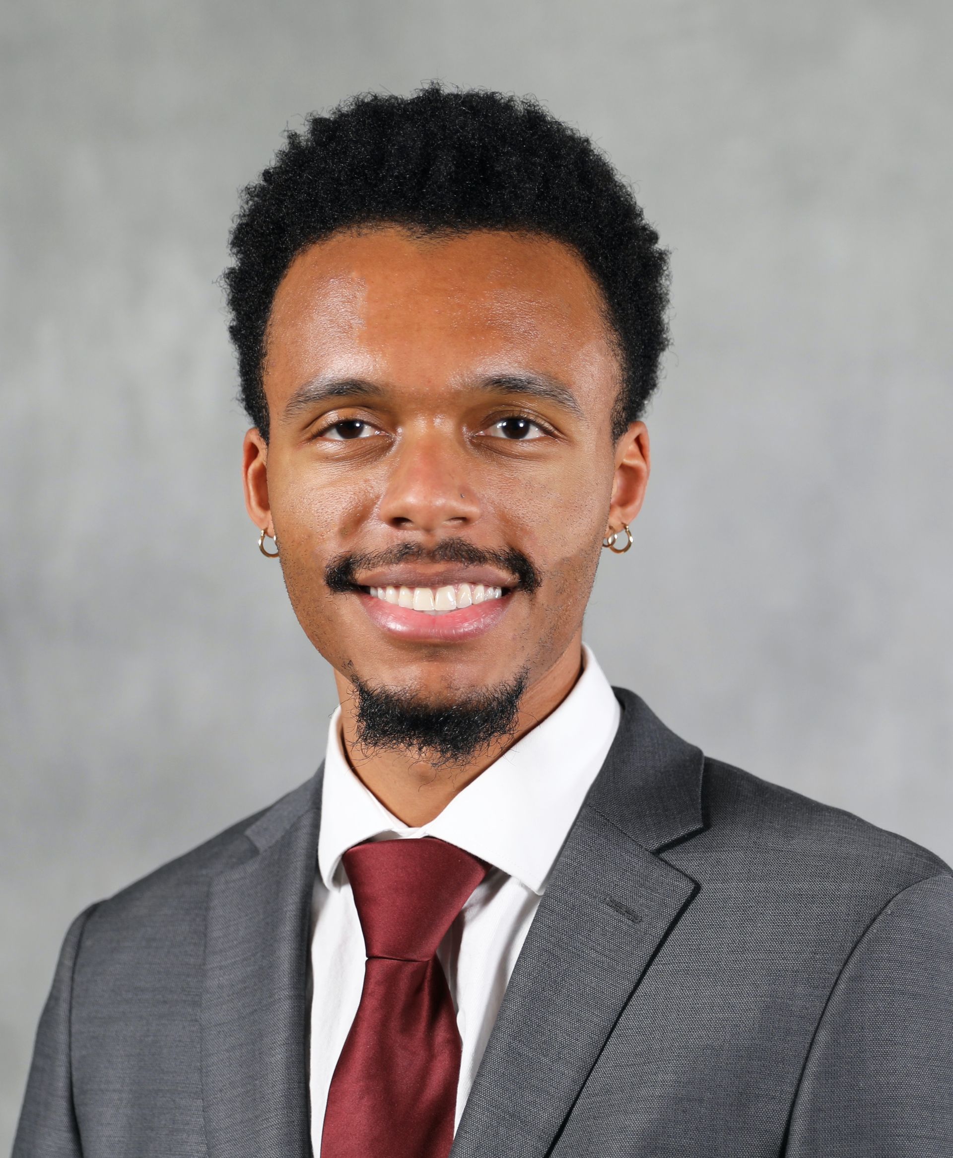 A smiling man wearing a gray suit, white collared shirt, and a dark red tie against a plain gray background.